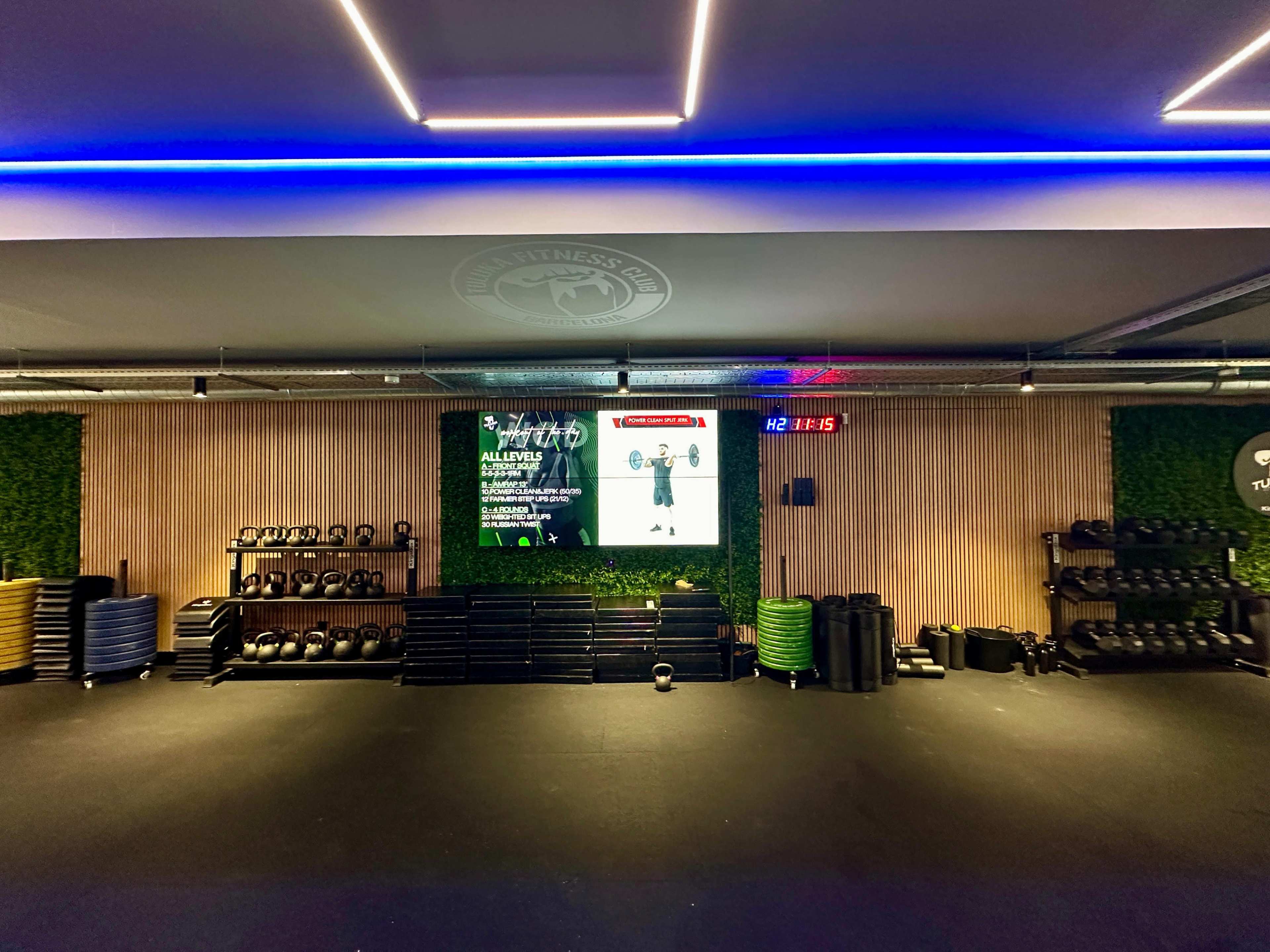 A gym interior with various weights, exercise mats, and a digital display showing a fitness class schedule on a wooden wall.