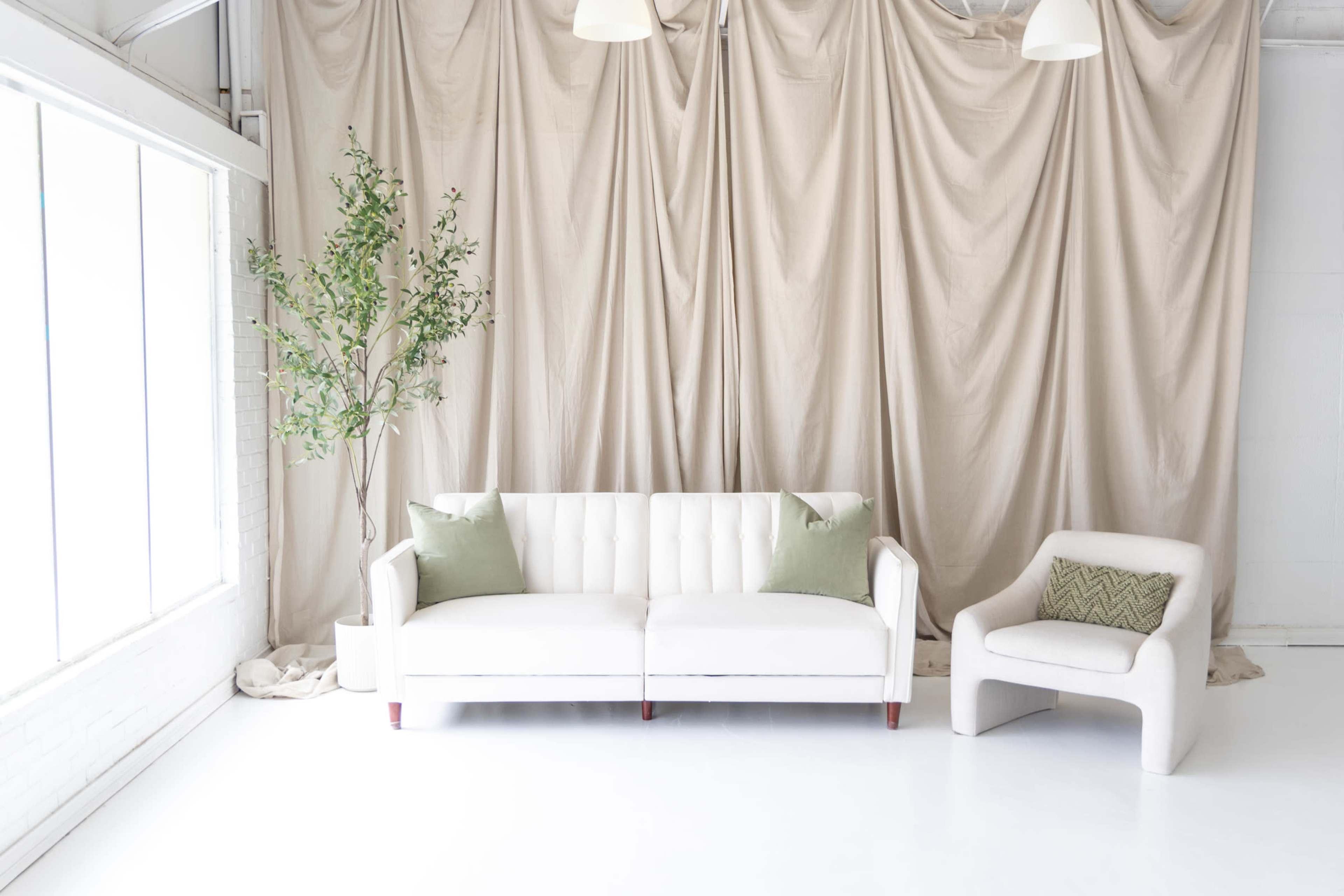 The image features a minimalist living room with a white sofa and an accent chair, complemented by a leafy plant and soft drapery in the background.