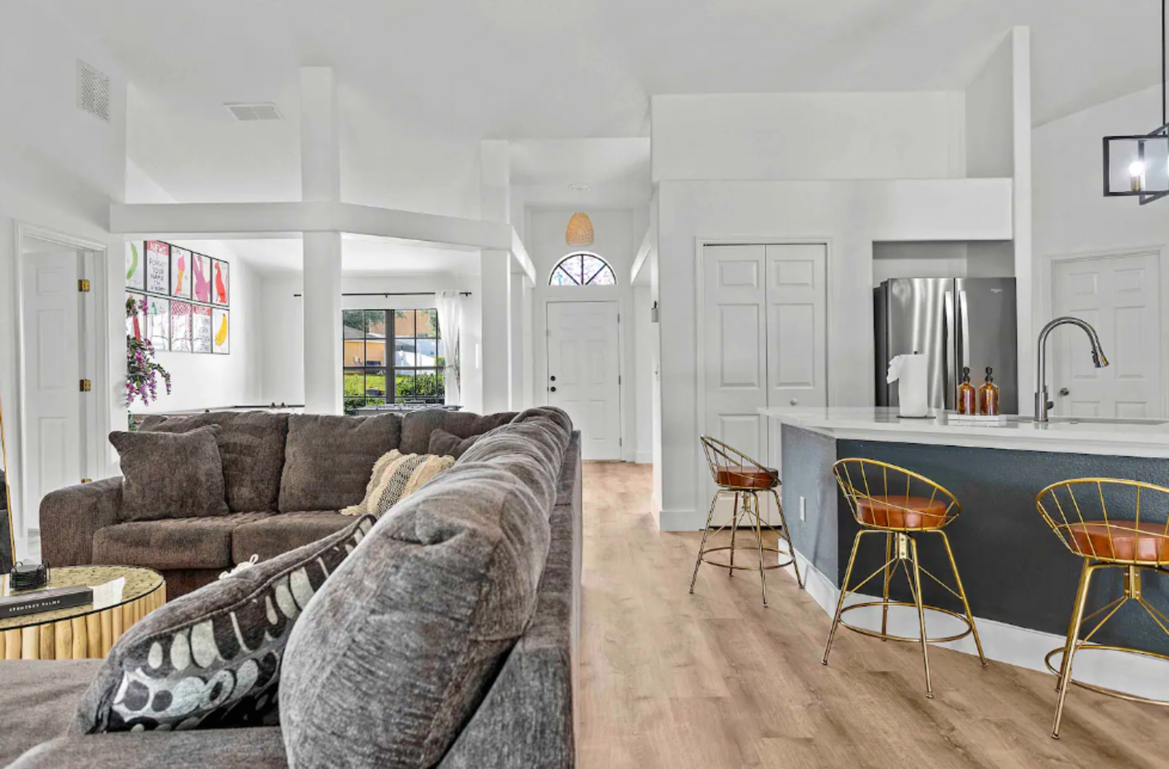 The image shows a modern living room with a large sectional sofa, an open layout leading to a kitchen area with bar stools and a refrigerator.