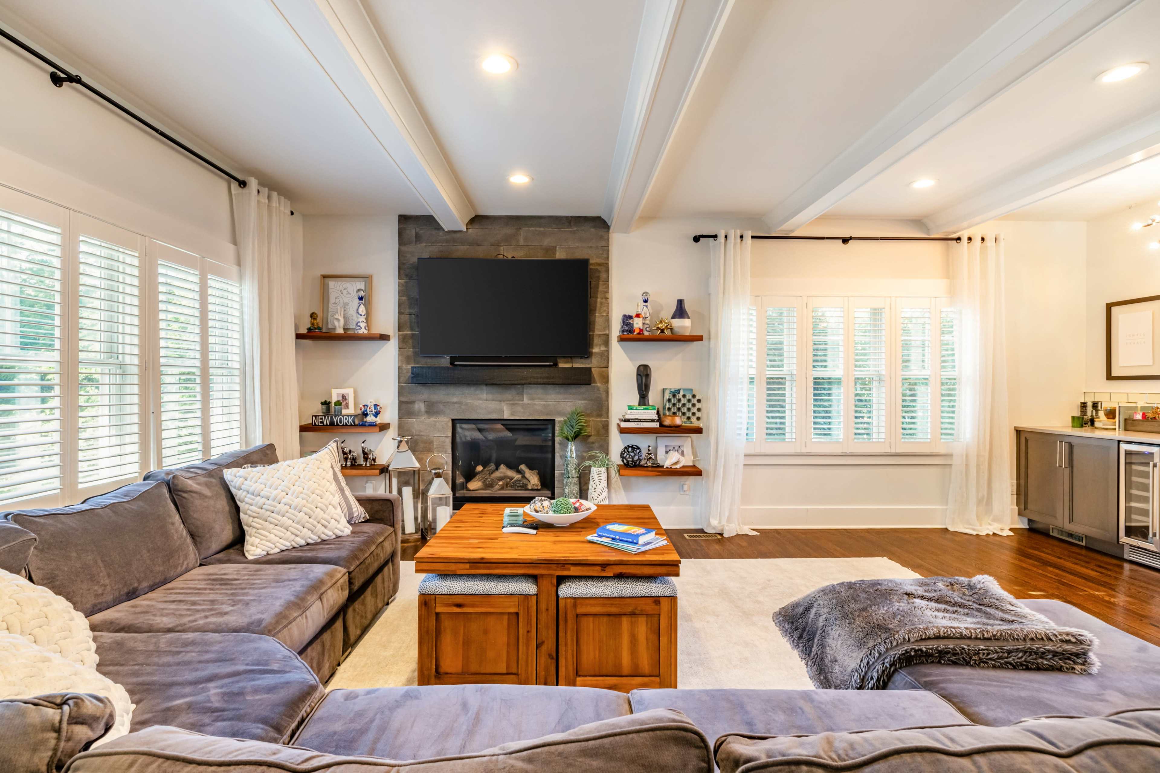 The image shows a cozy living room featuring a sectional sofa, a wooden coffee table, a stone fireplace with a television mounted above it, and large windows with sheer curtains.
