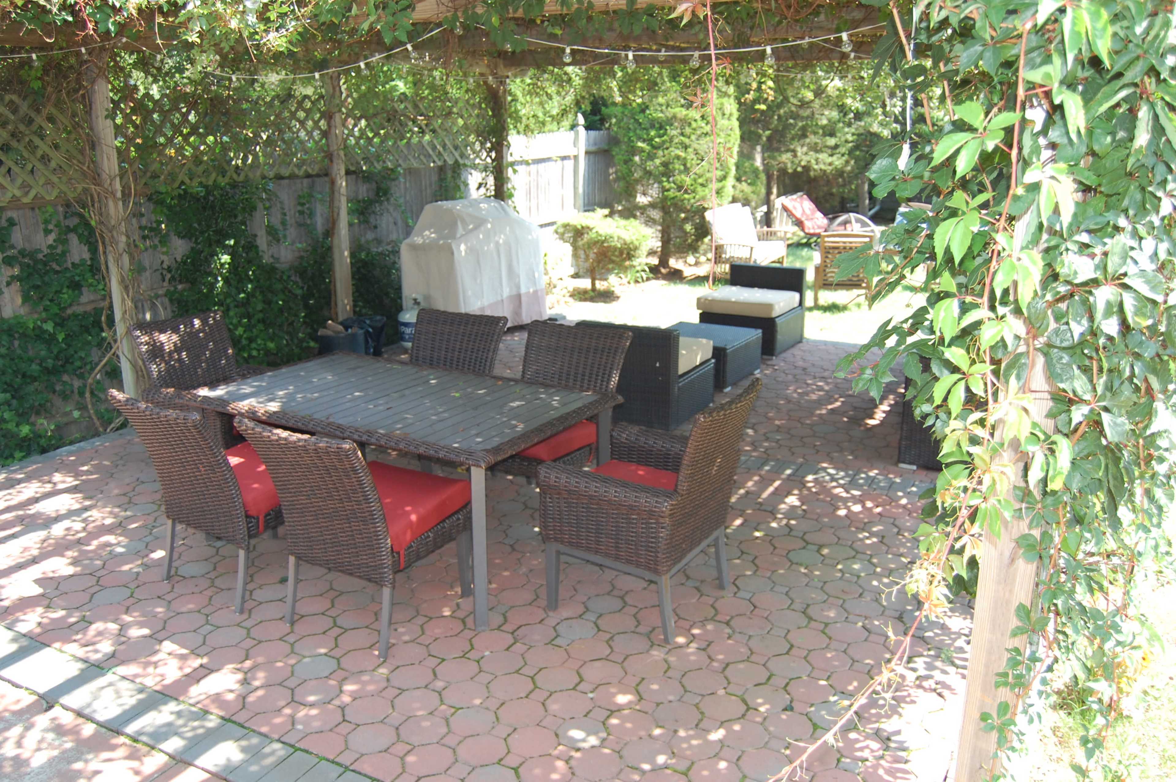 A patio features a wooden table surrounded by eight wicker chairs with red cushions, under a covered area with various seating arrangements in the background.