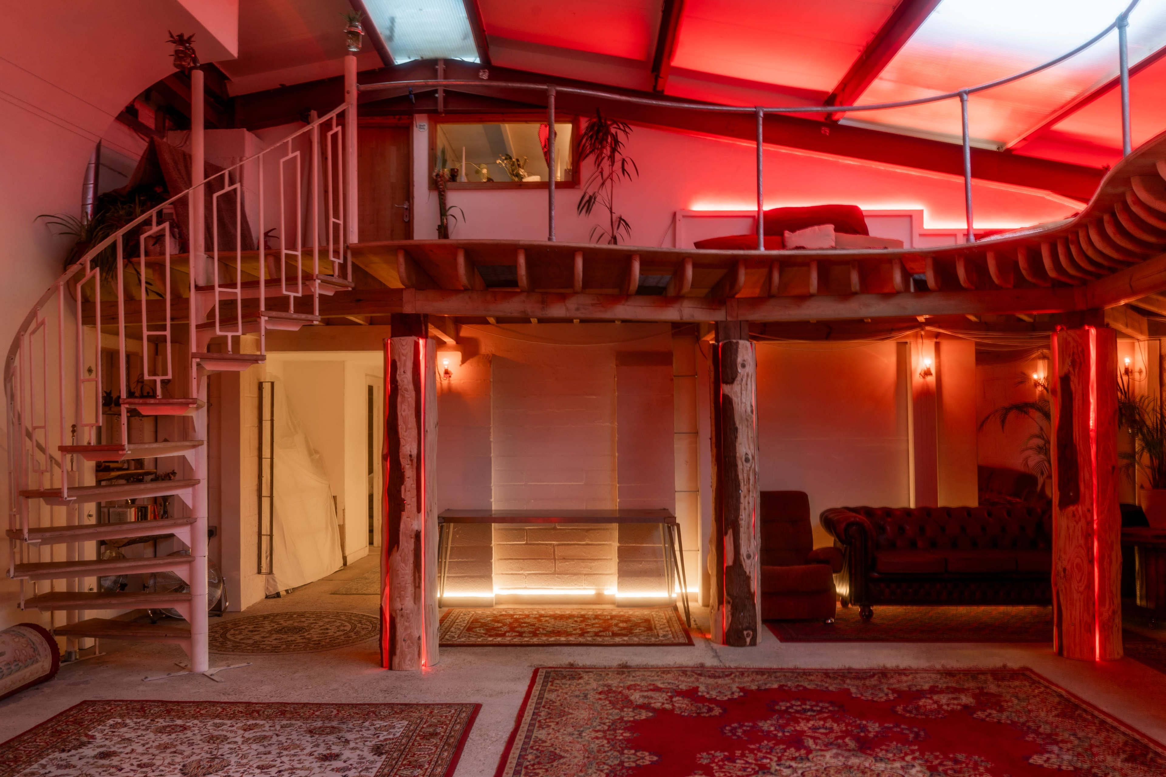 The image shows a room with a spiral staircase, wooden beams, and red ambient lighting, featuring a cozy seating area and decorative rugs.