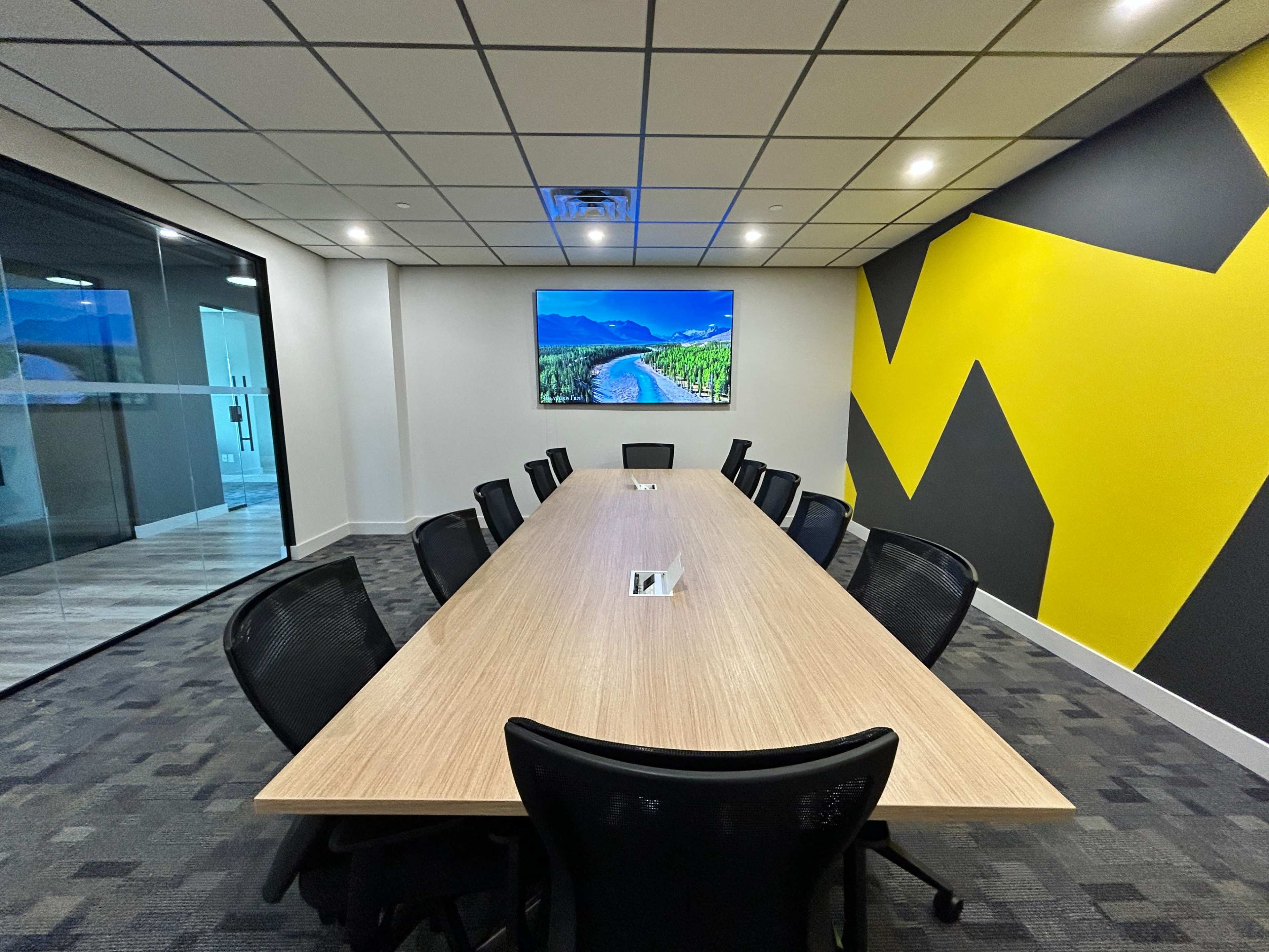 A modern conference room with a long wooden table surrounded by black chairs and a large screen displaying a landscape on the wall.