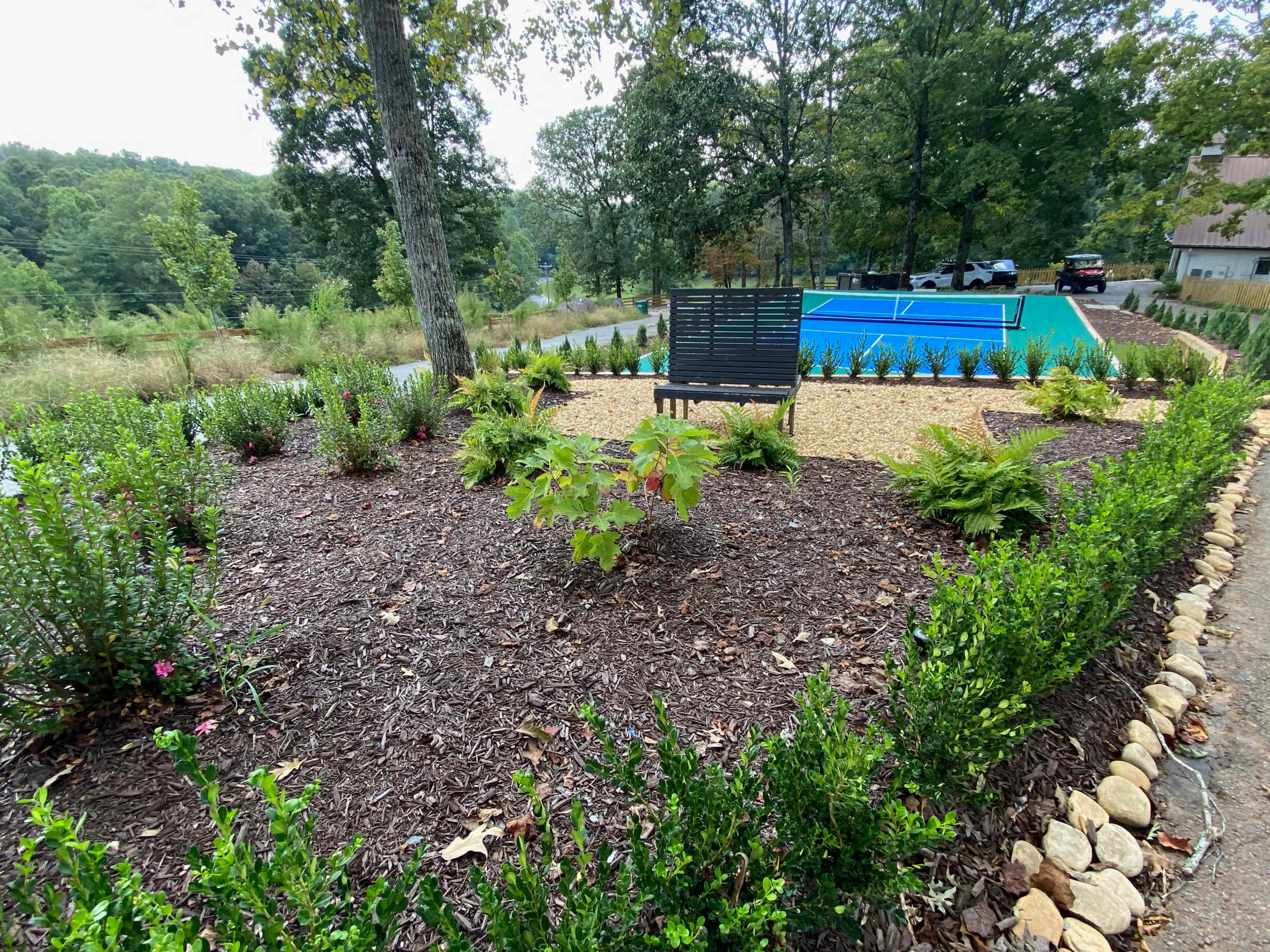 A landscaped area featuring a bench surrounded by plants and mulch, adjacent to a blue tennis court in a wooded setting.