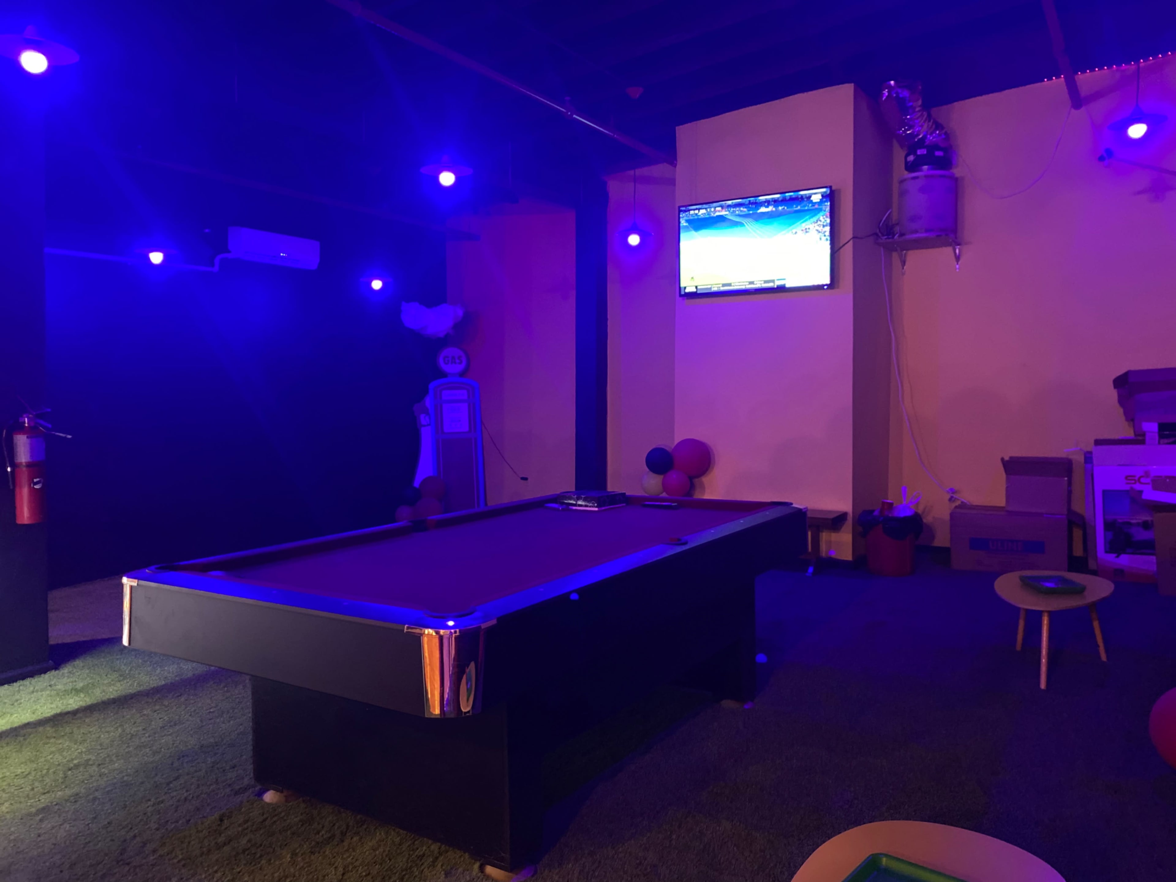 A billiard table in a dimly lit room with a television displaying a game and colorful lights.