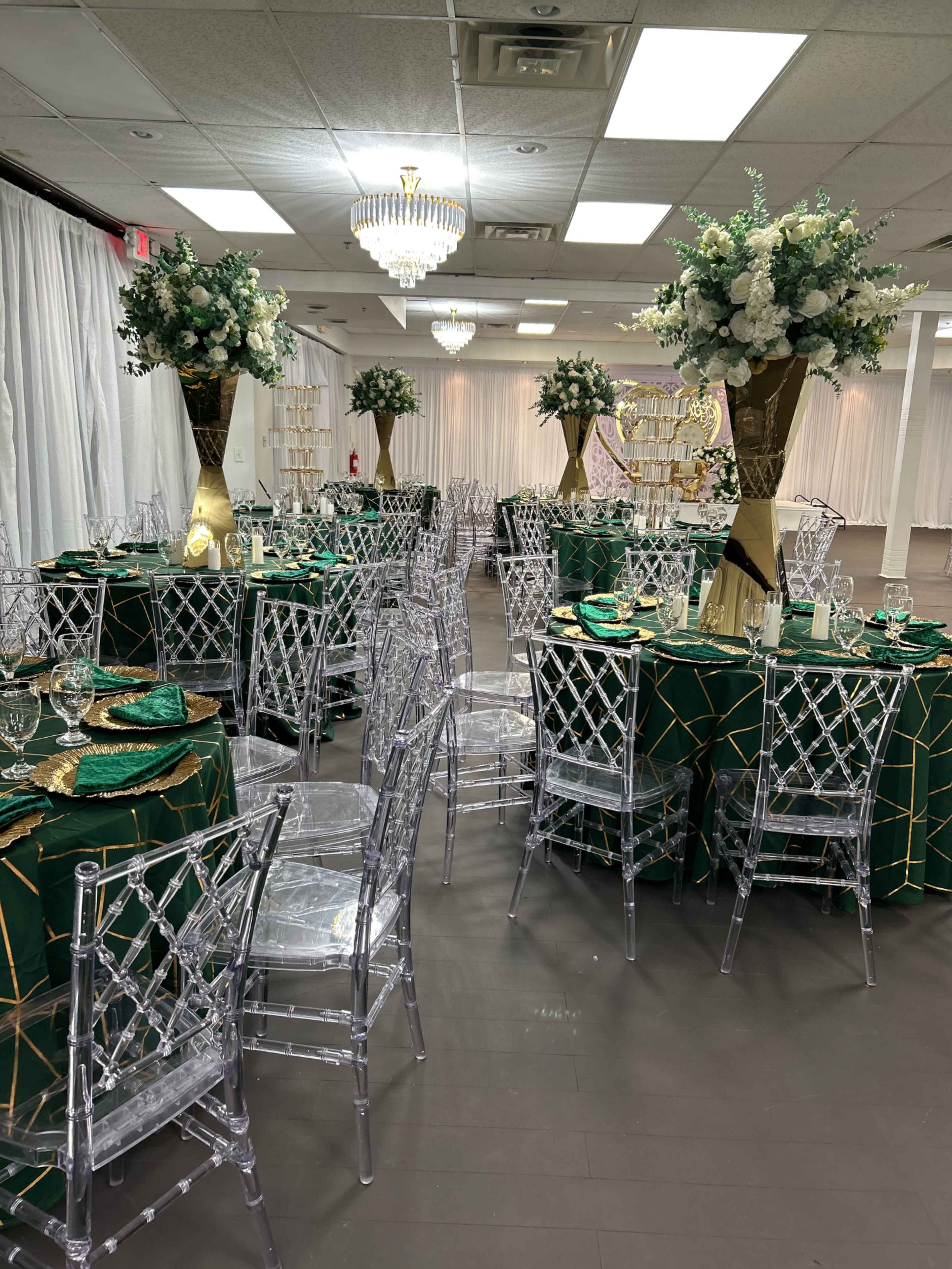 The image shows a dining area set for an event, featuring round tables with green tablecloths, gold accents, clear acrylic chairs, and floral centerpieces.