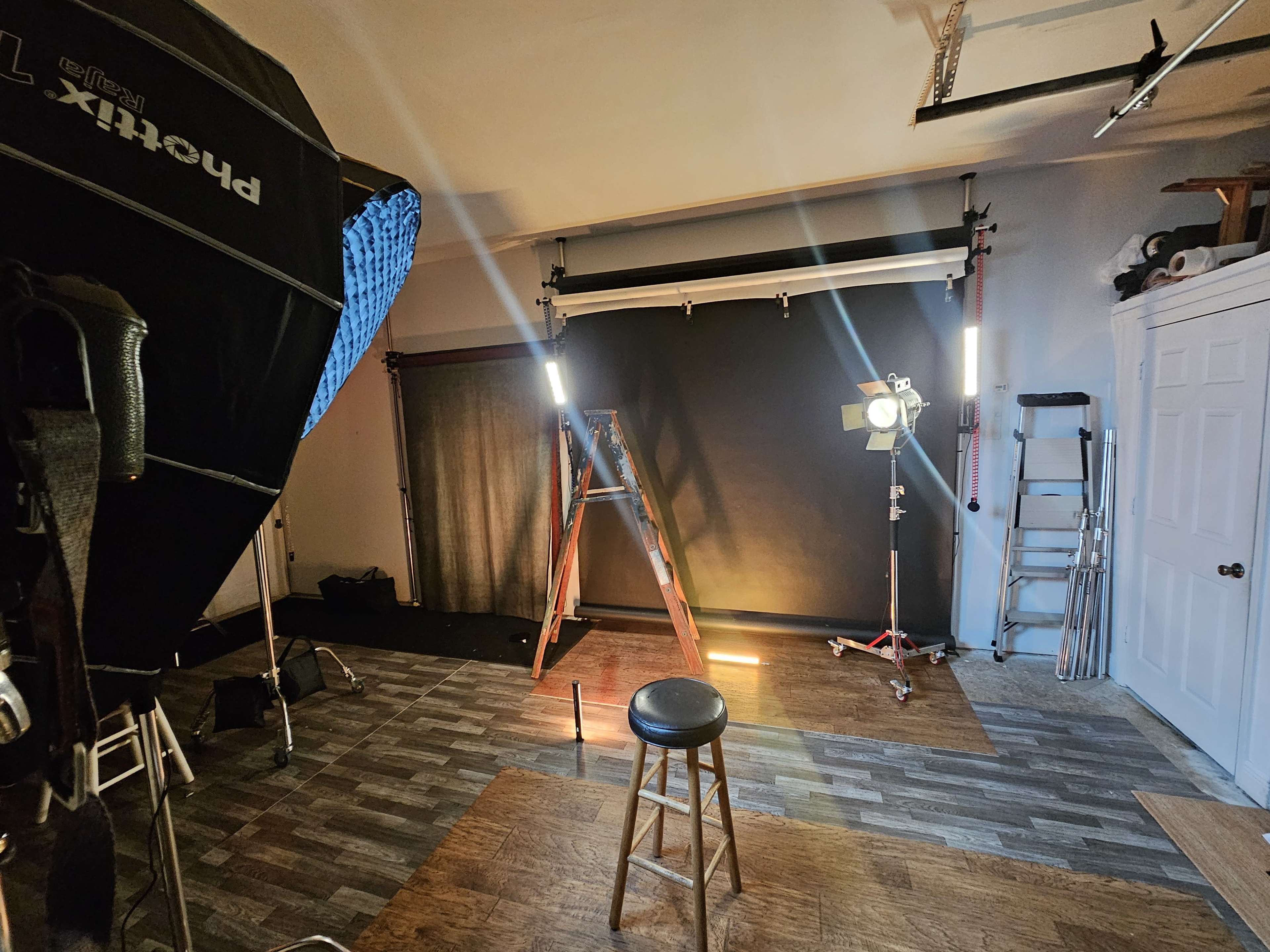 The image shows a photography studio setup with a black backdrop, a stool, lights, and equipment positioned around the area.