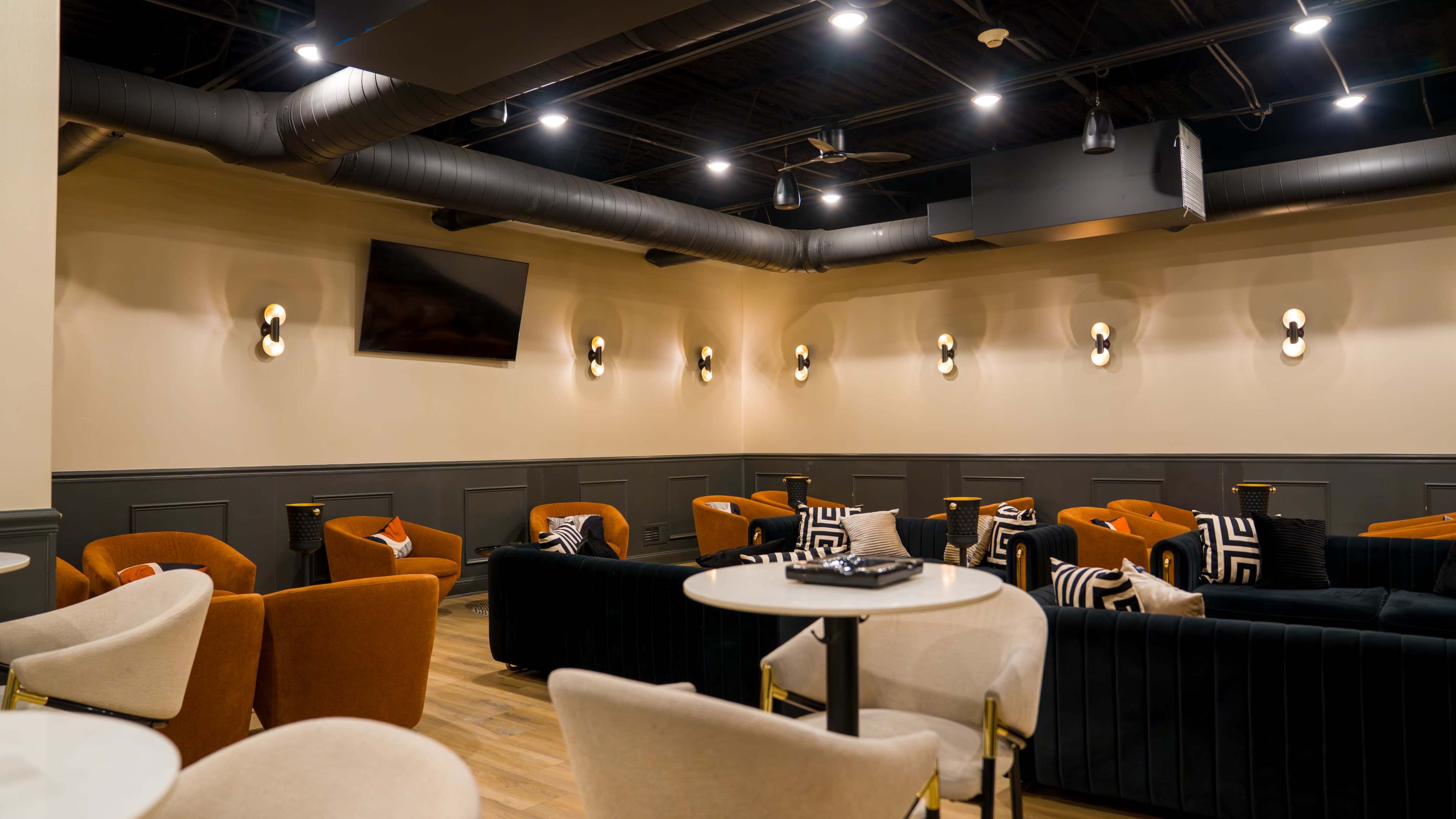 The image shows a modern lounge area with various seating options, including orange and dark green chairs, and several wall-mounted lights, all under a black ceiling with visible ductwork.