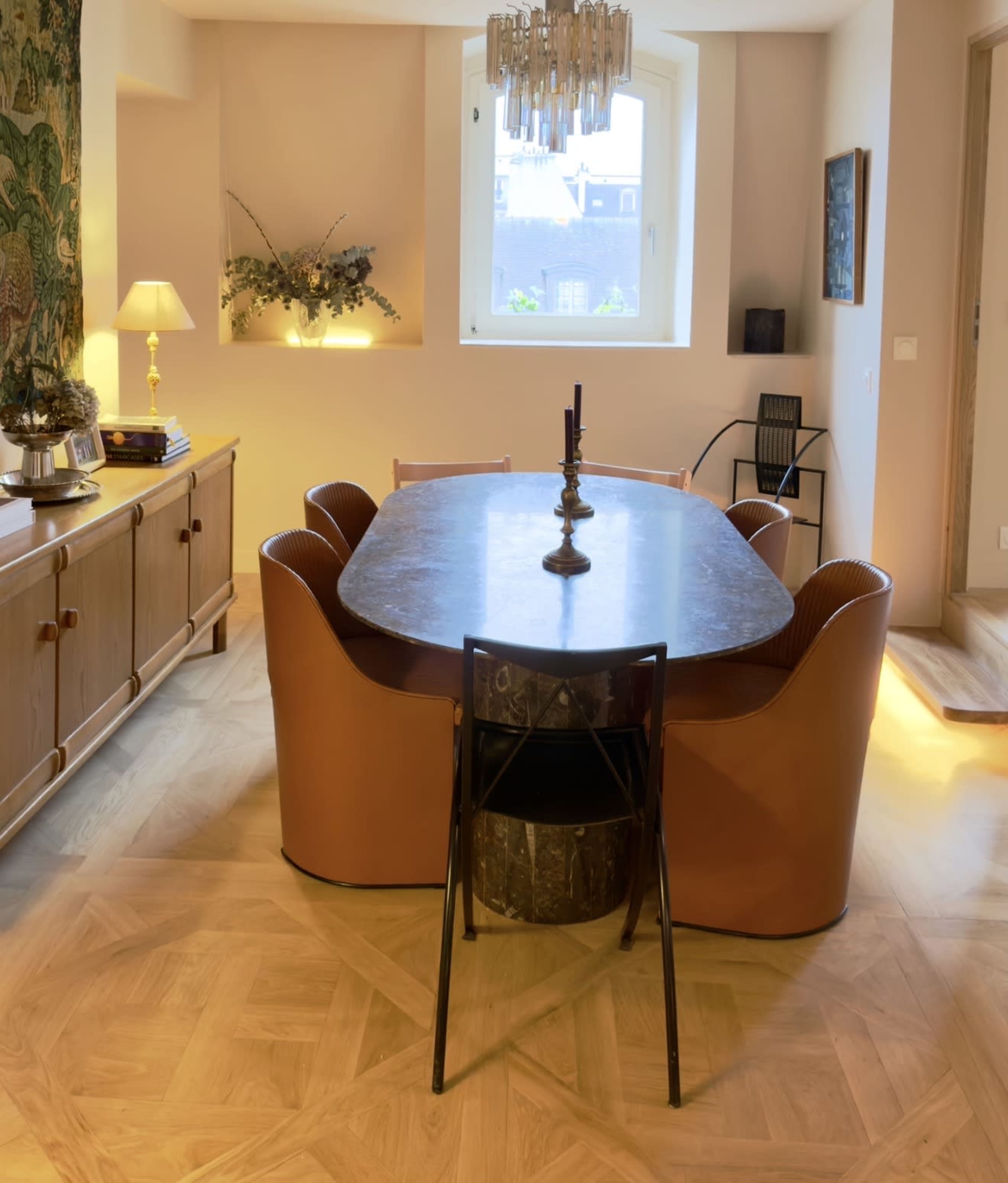 The image shows a modern dining room featuring an oval table surrounded by leather chairs, with a large window and a chandelier overhead.