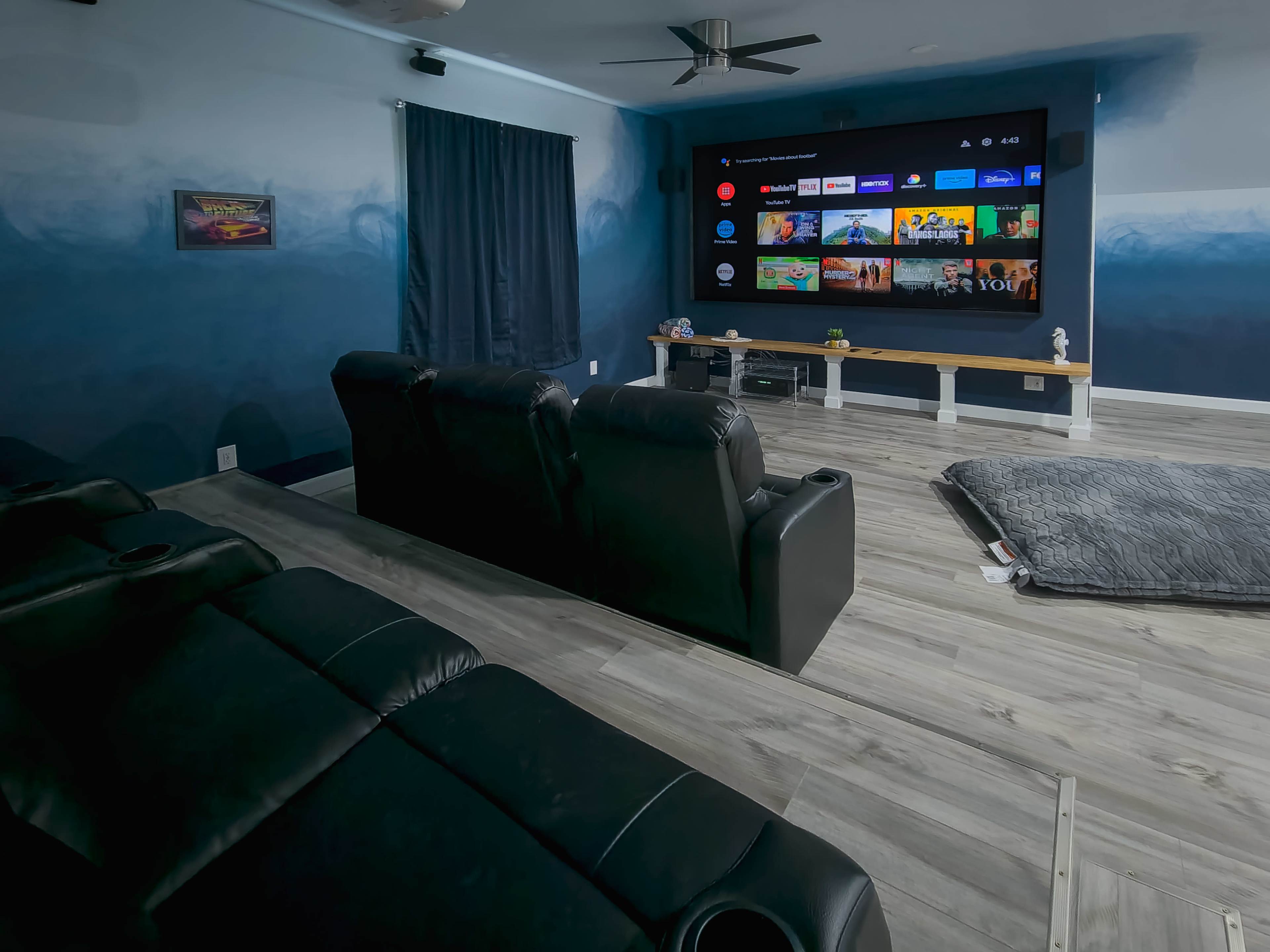 A home theater room features black reclining chairs facing a large wall-mounted screen, with a light-colored wooden shelf underneath, and a gray area rug on the floor.