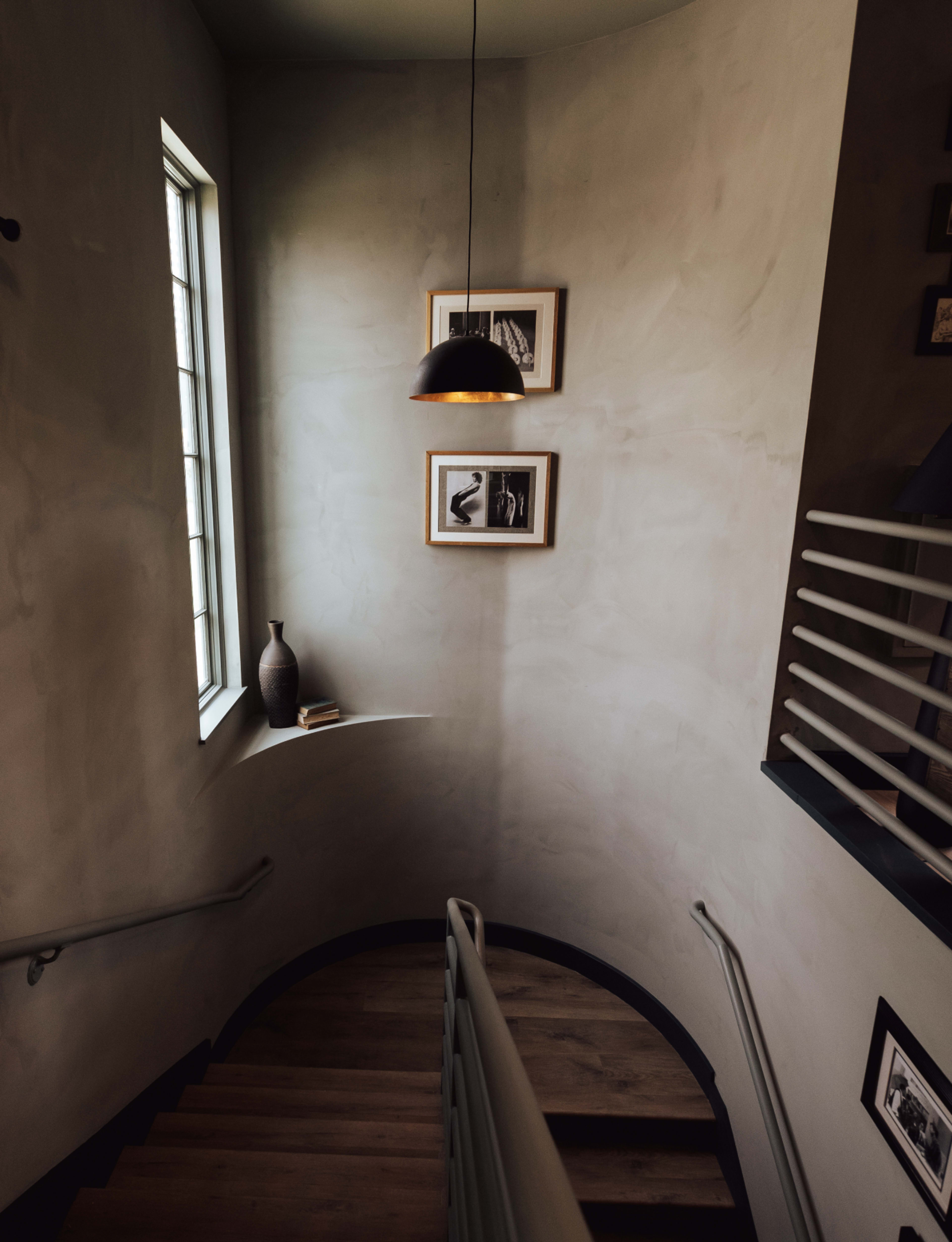 A curved staircase descends into a softly lit area featuring a window, framed images, and a decorative vase.