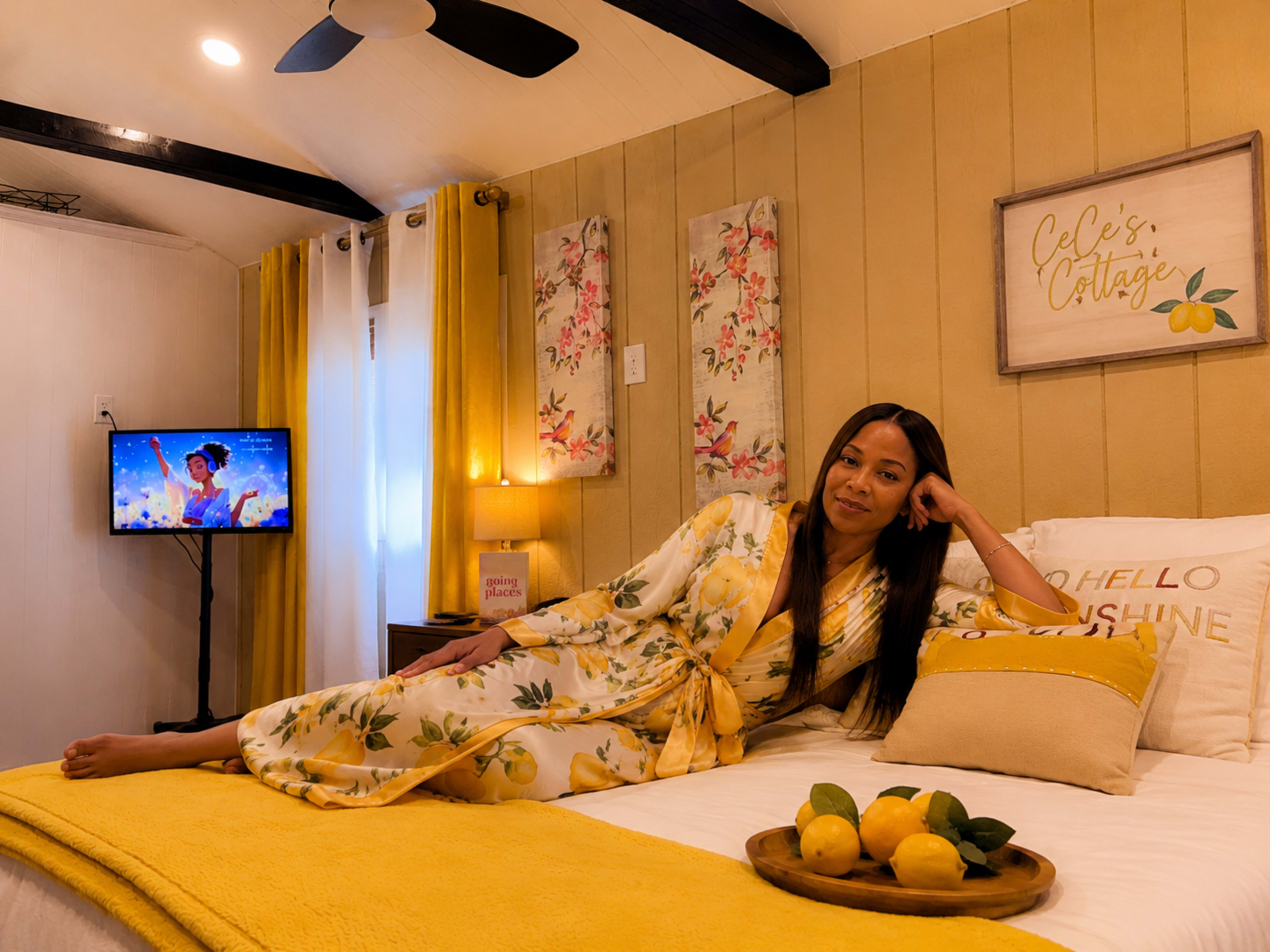 A woman in a floral robe lounges on a white bed adorned with yellow accents in a cozy bedroom with lemon-themed decor and a television in the background.