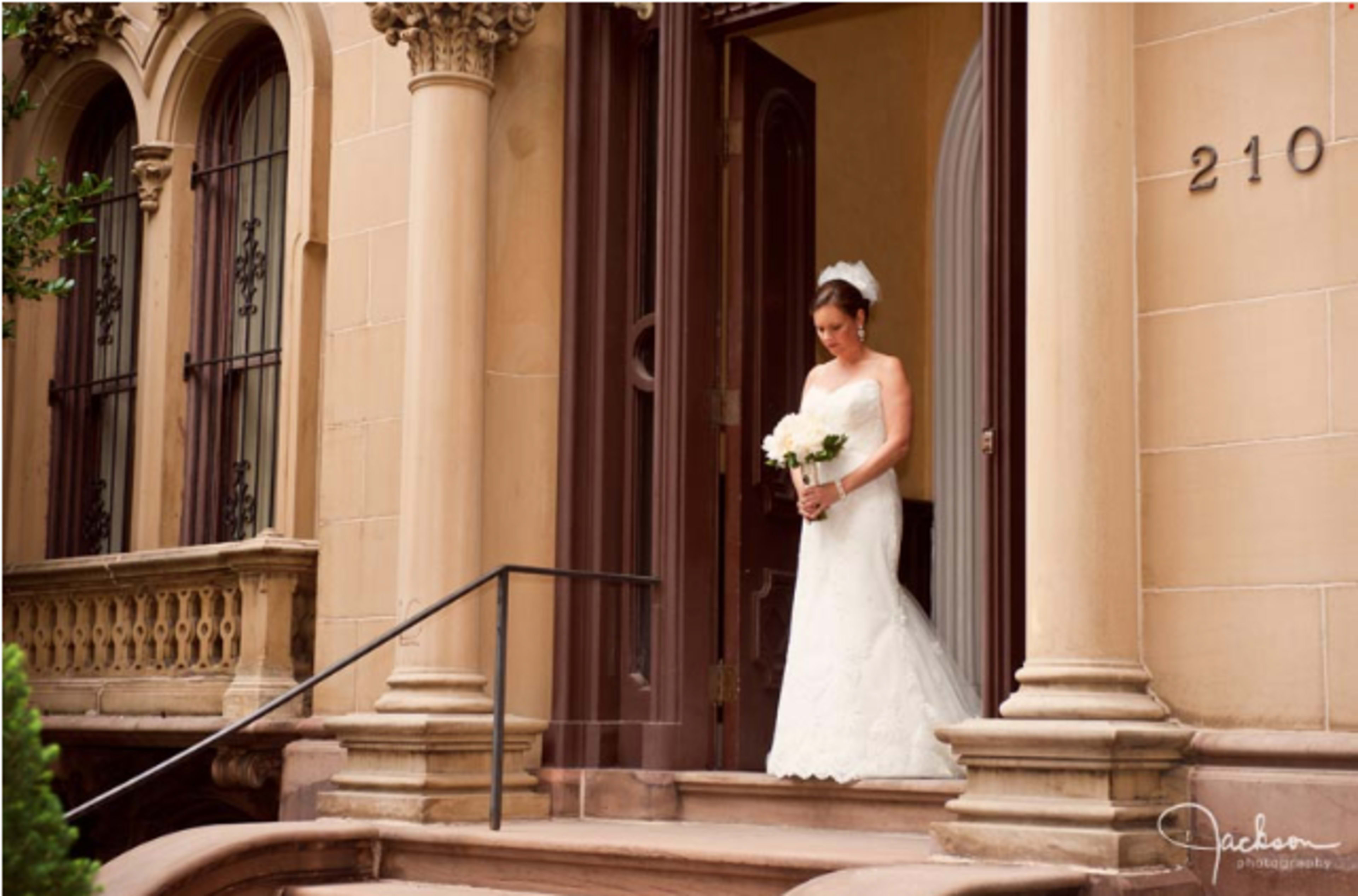 A bride in a white gown stands on the steps of a grand building, holding a bouquet of white flowers.