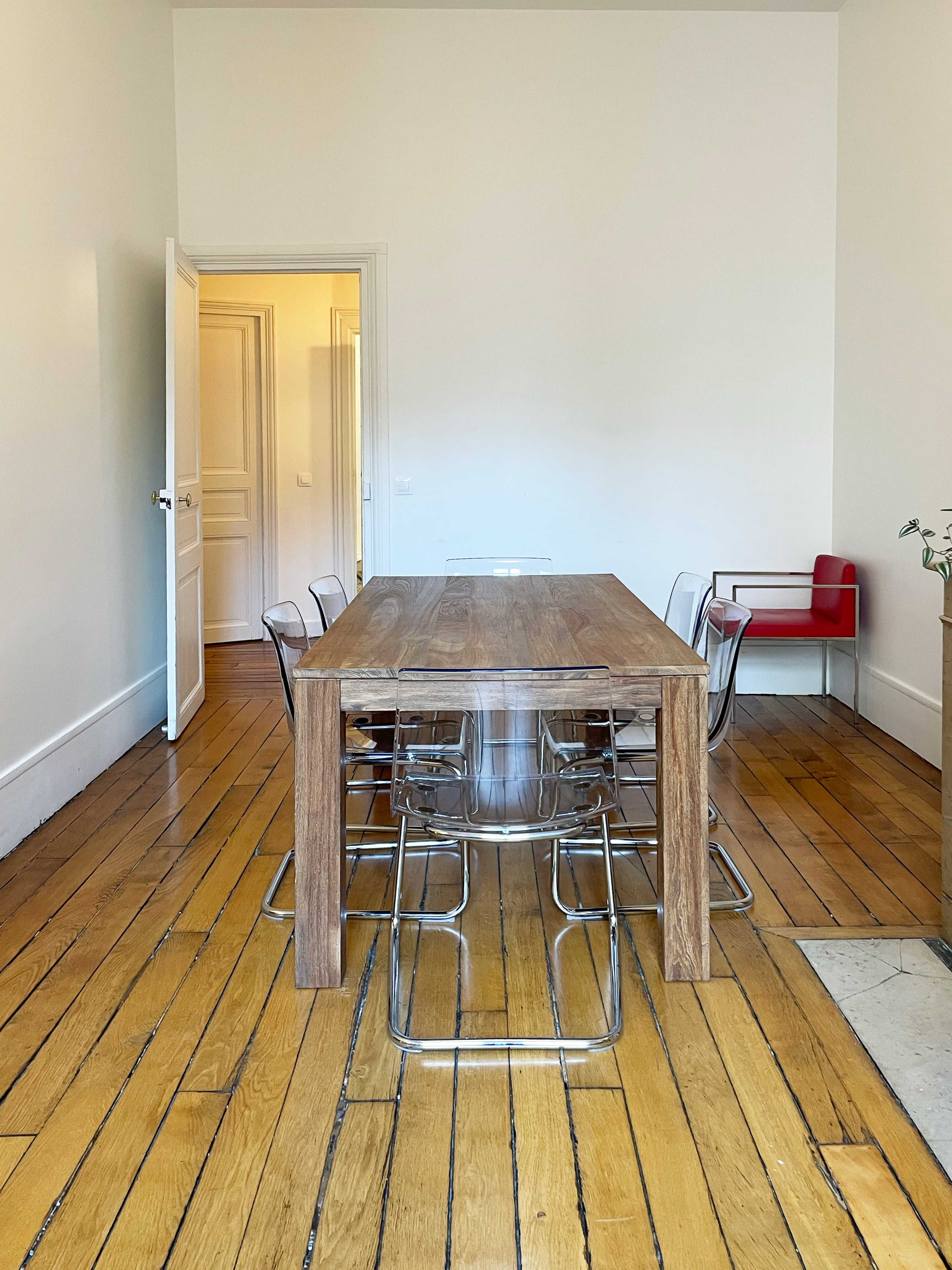 A wooden dining table is centered in a room with hardwood floors, surrounded by clear acrylic chairs, and a red chair is visible against the wall.