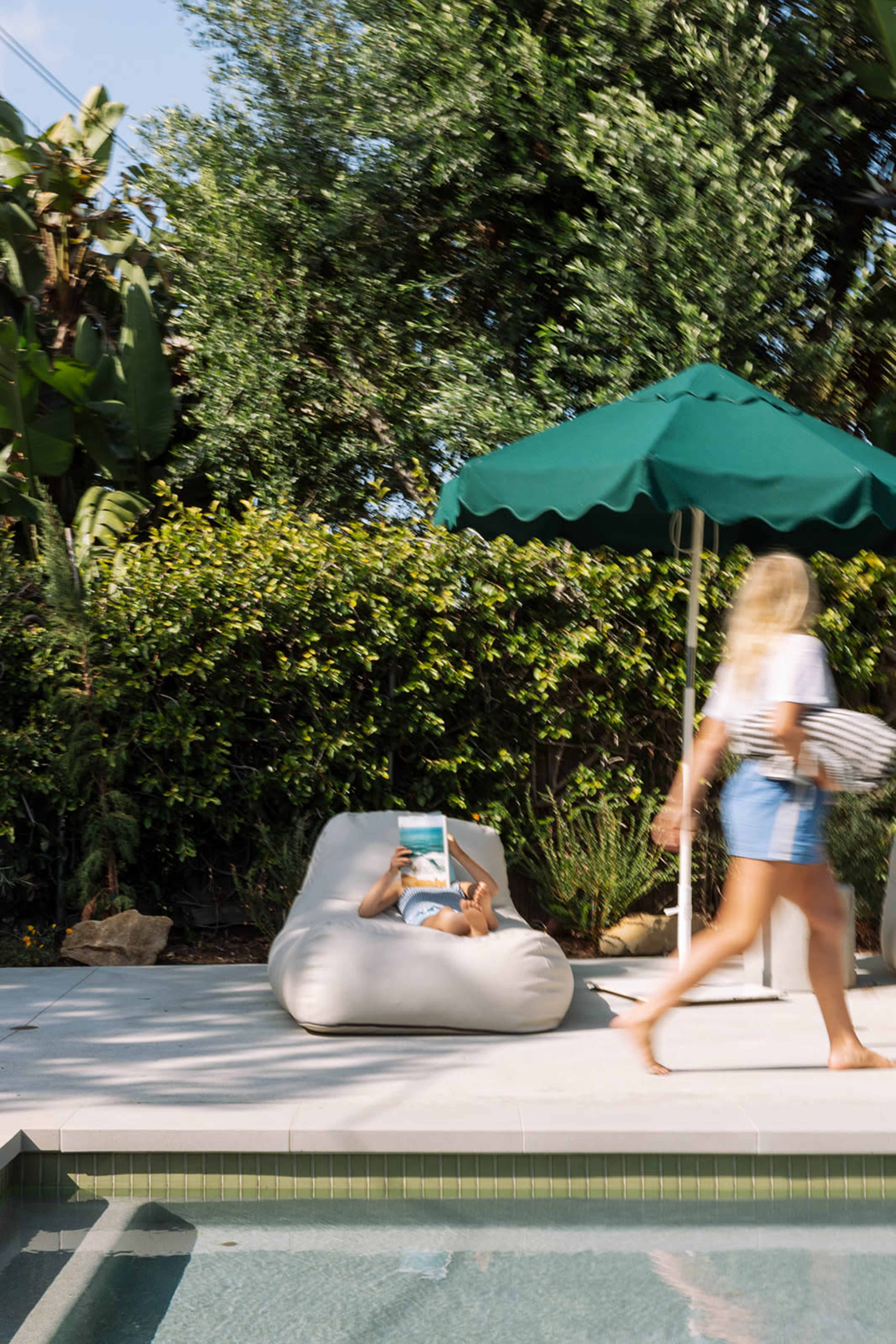 A person lounges on a bean bag chair by a pool while another figure walks past under a green umbrella.