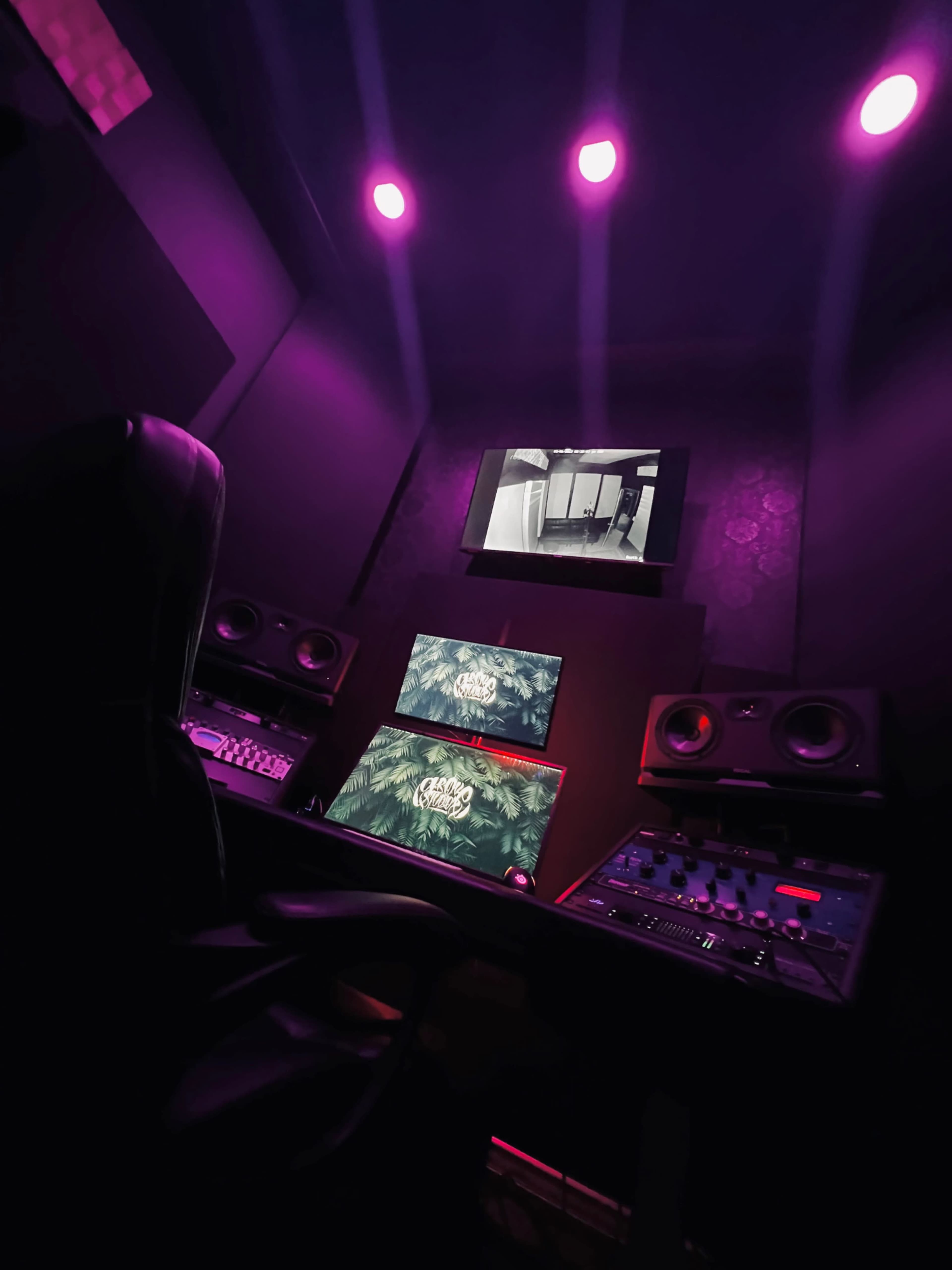 A dimly lit music studio features two monitors, audio equipment, and colorful lighting.