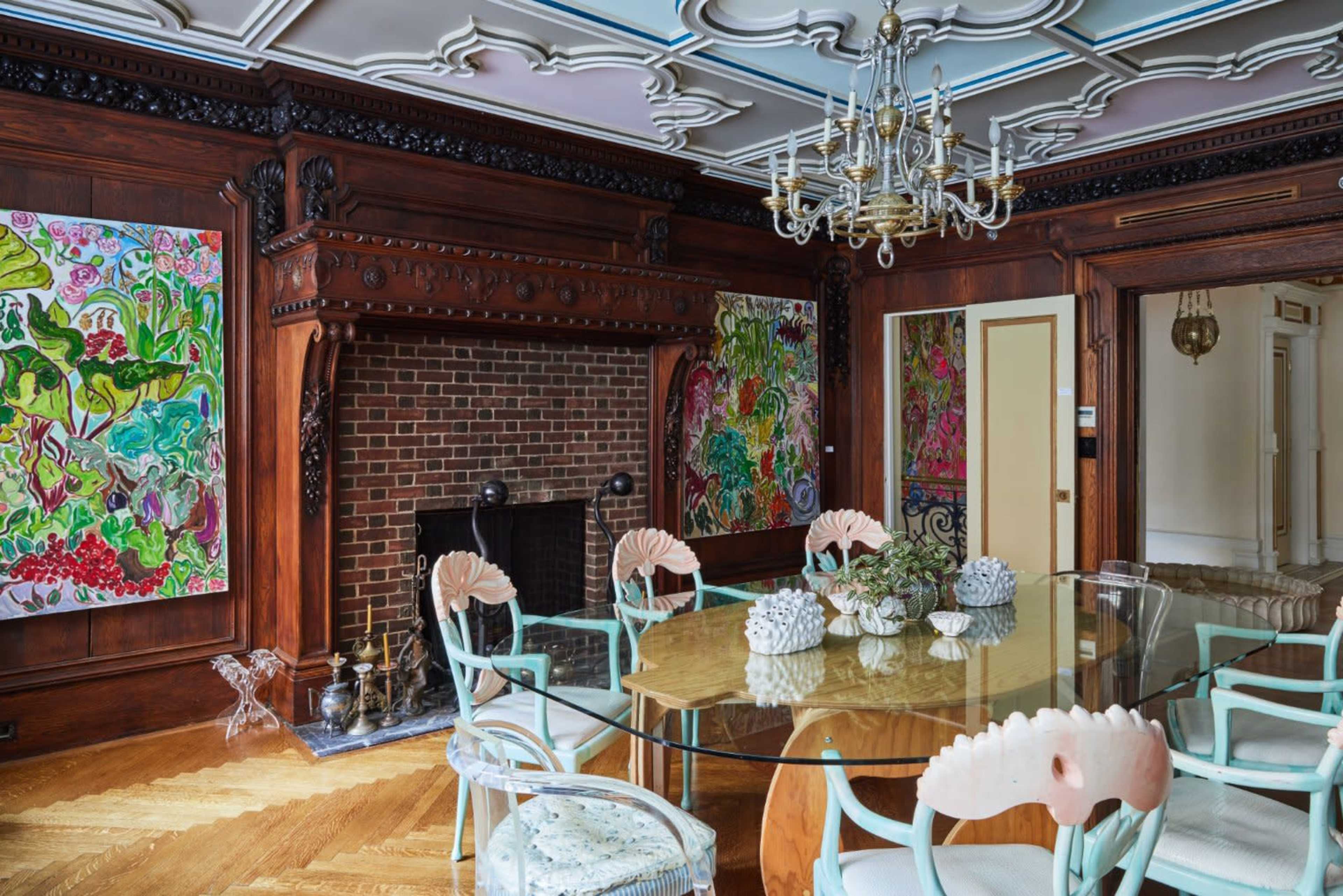 The image shows an elegant dining room featuring a round glass table surrounded by ornate chairs, with colorful artwork on the walls and a brick fireplace.