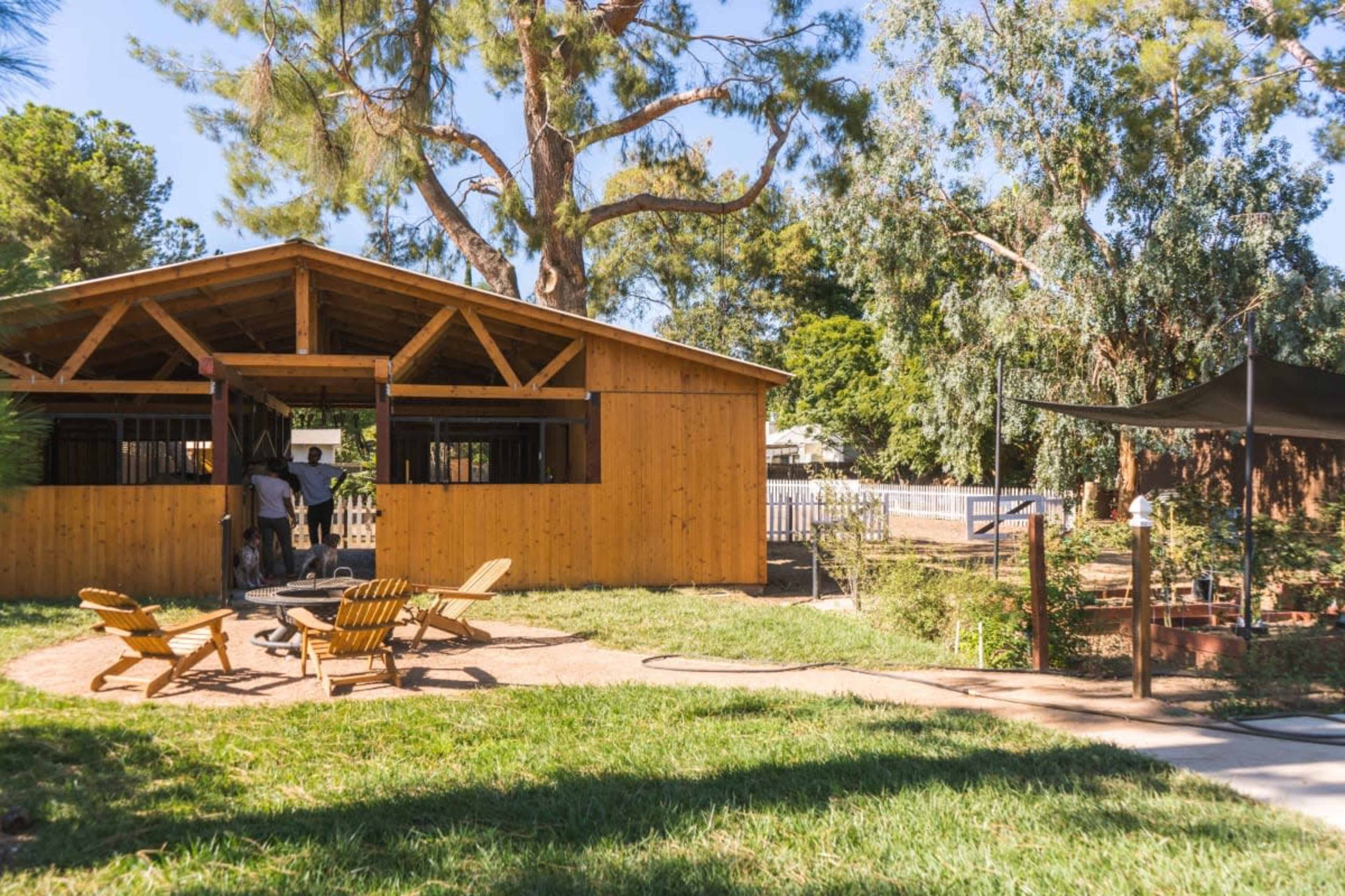 A wooden structure with a fenced yard, surrounded by trees, features two Adirondack chairs and a fire pit in the foreground.