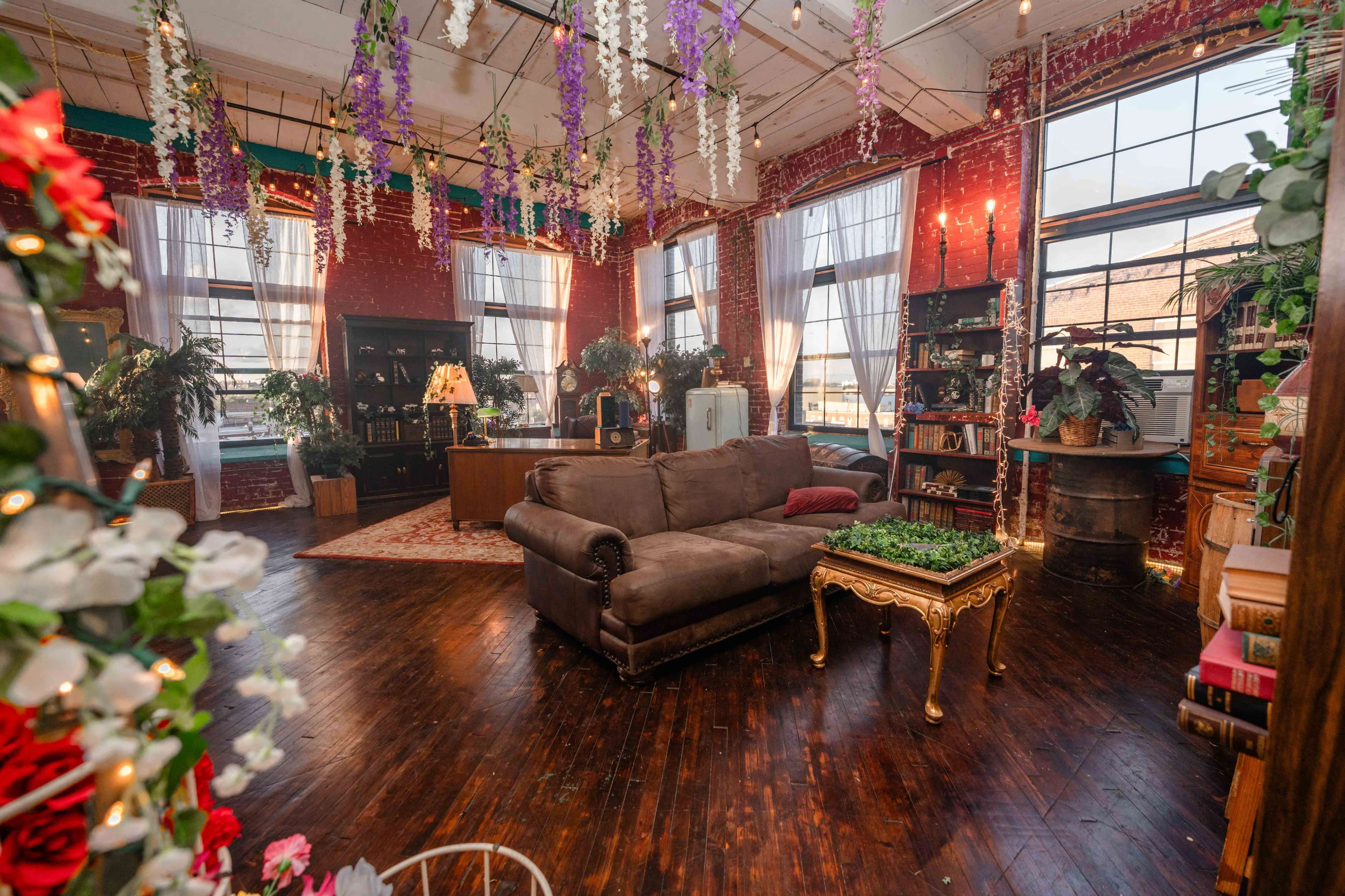 The image shows a spacious, well-lit room with large windows, a brown sofa, and various plants, decorated with hanging flowers and bookshelves.