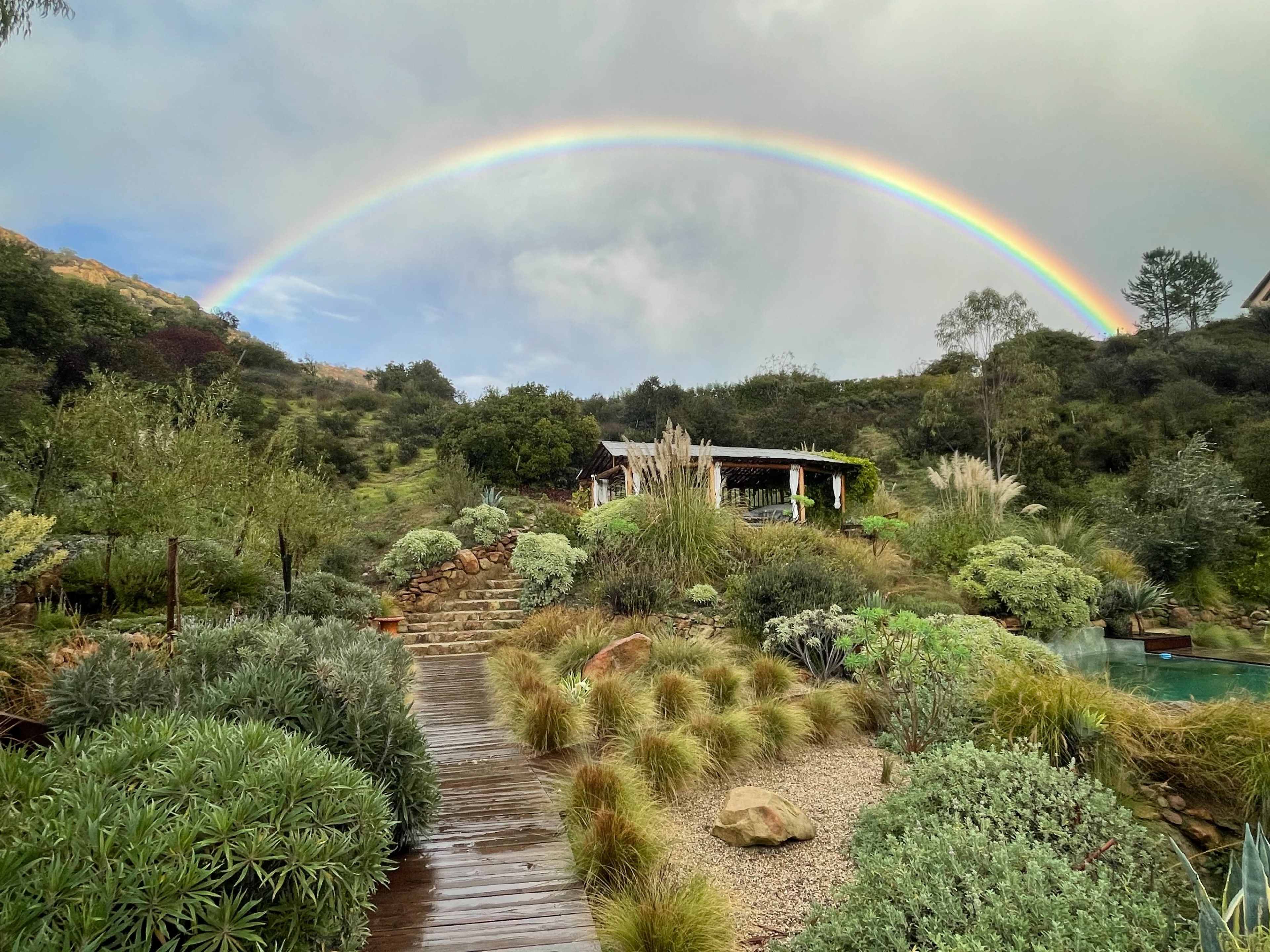 A vibrant rainbow arcs over a lush, landscaped garden with a pathway leading to a rustic structure amidst greenery and rocks.