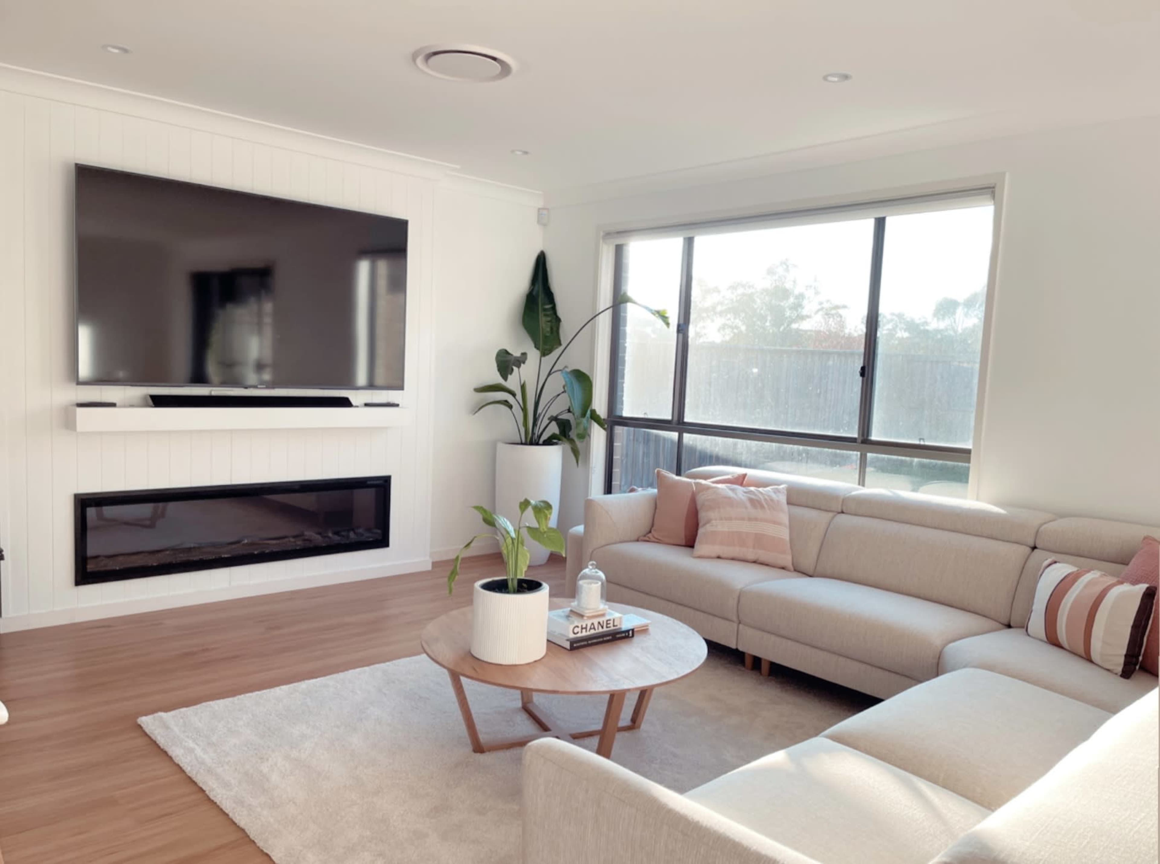 A modern living room features a large wall-mounted television above a linear electric fireplace, with a sectional sofa, a round coffee table, and a window offering natural light.