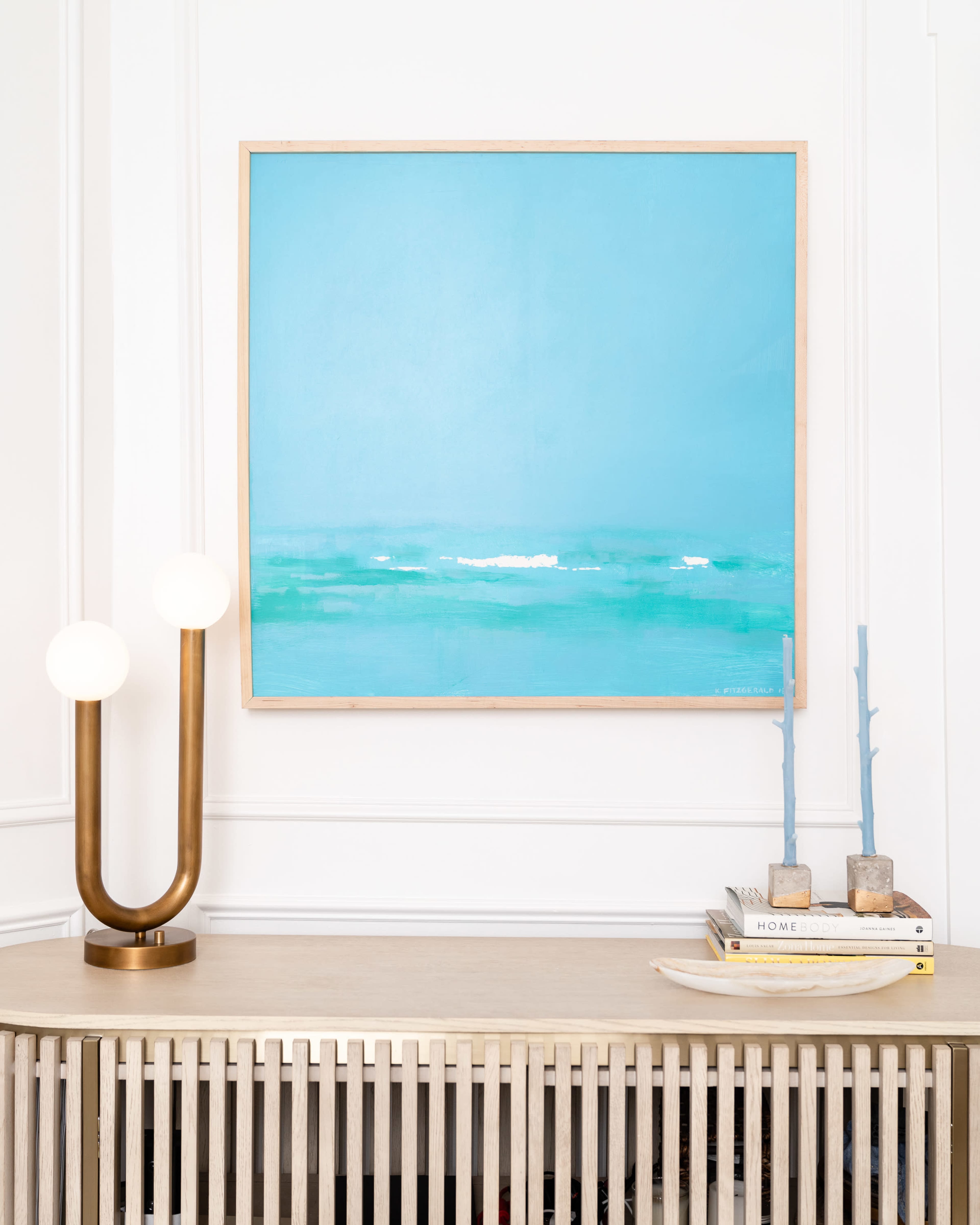 A blue abstract painting hangs on a white wall above a light wood side table with a lamp and stacked books.
