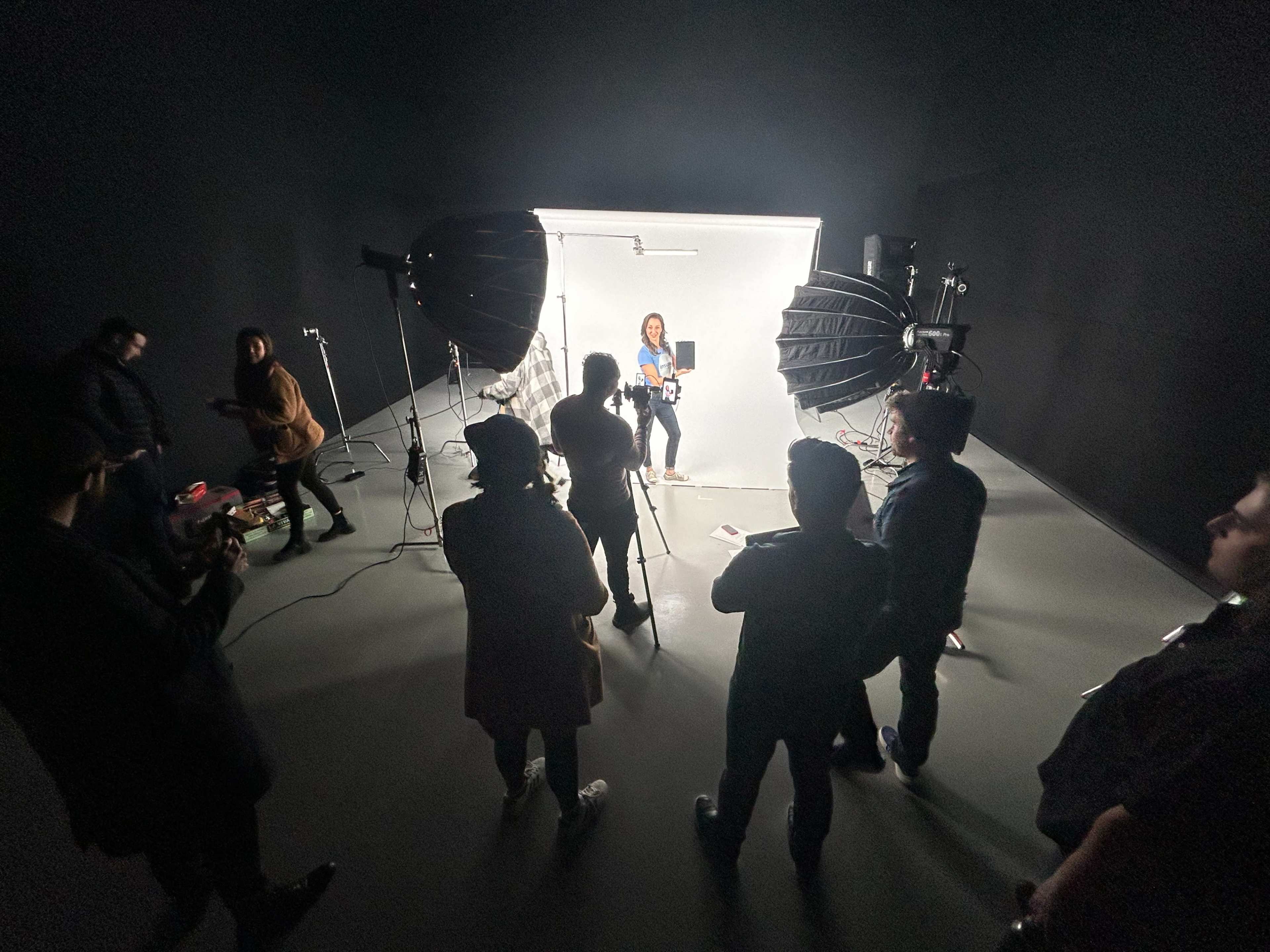 A group of people surrounds a person sitting in front of bright studio lights in a dark studio space.