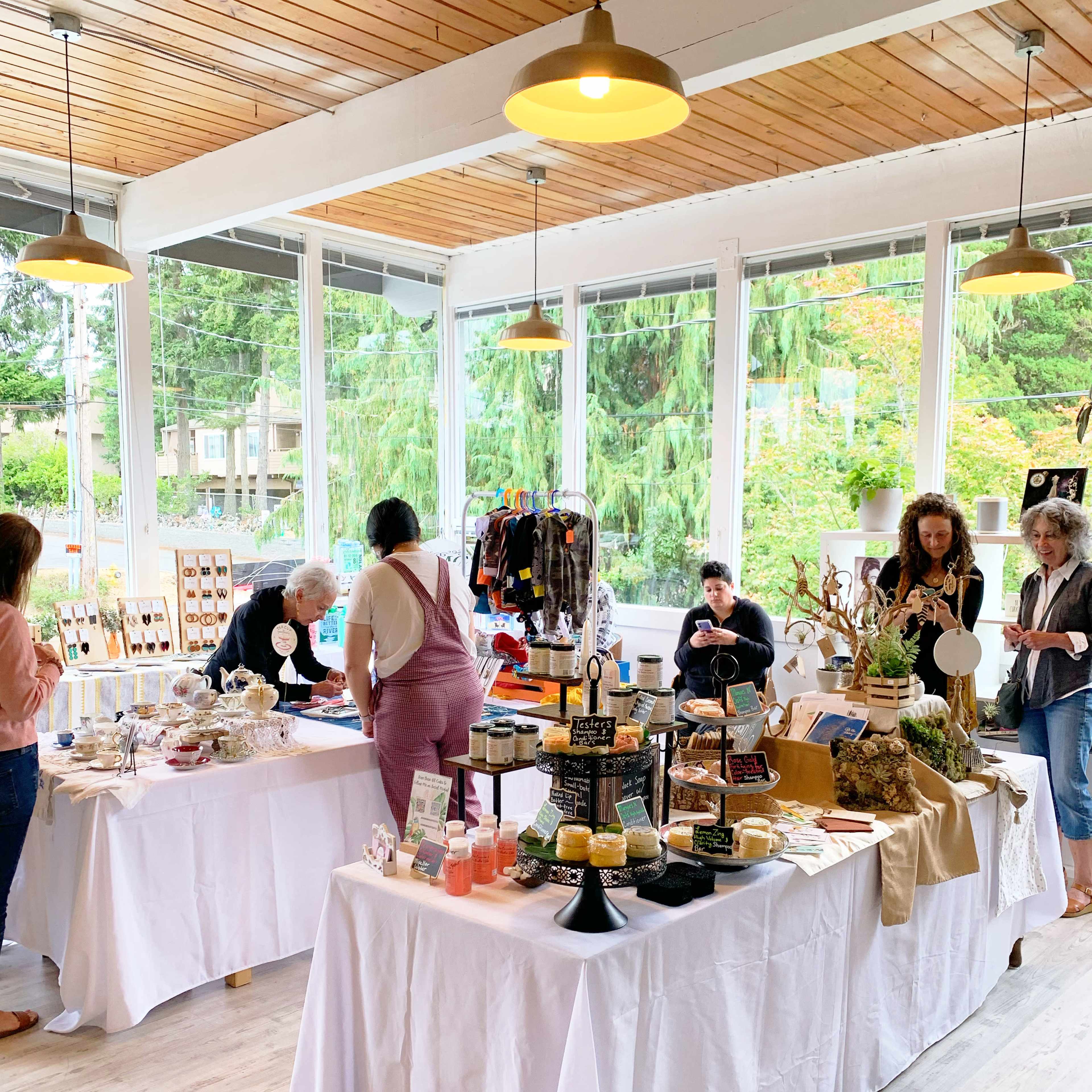 The scene shows a vibrant, indoor market with several tables displaying various handmade goods and food items, while customers browse and interact with vendors.