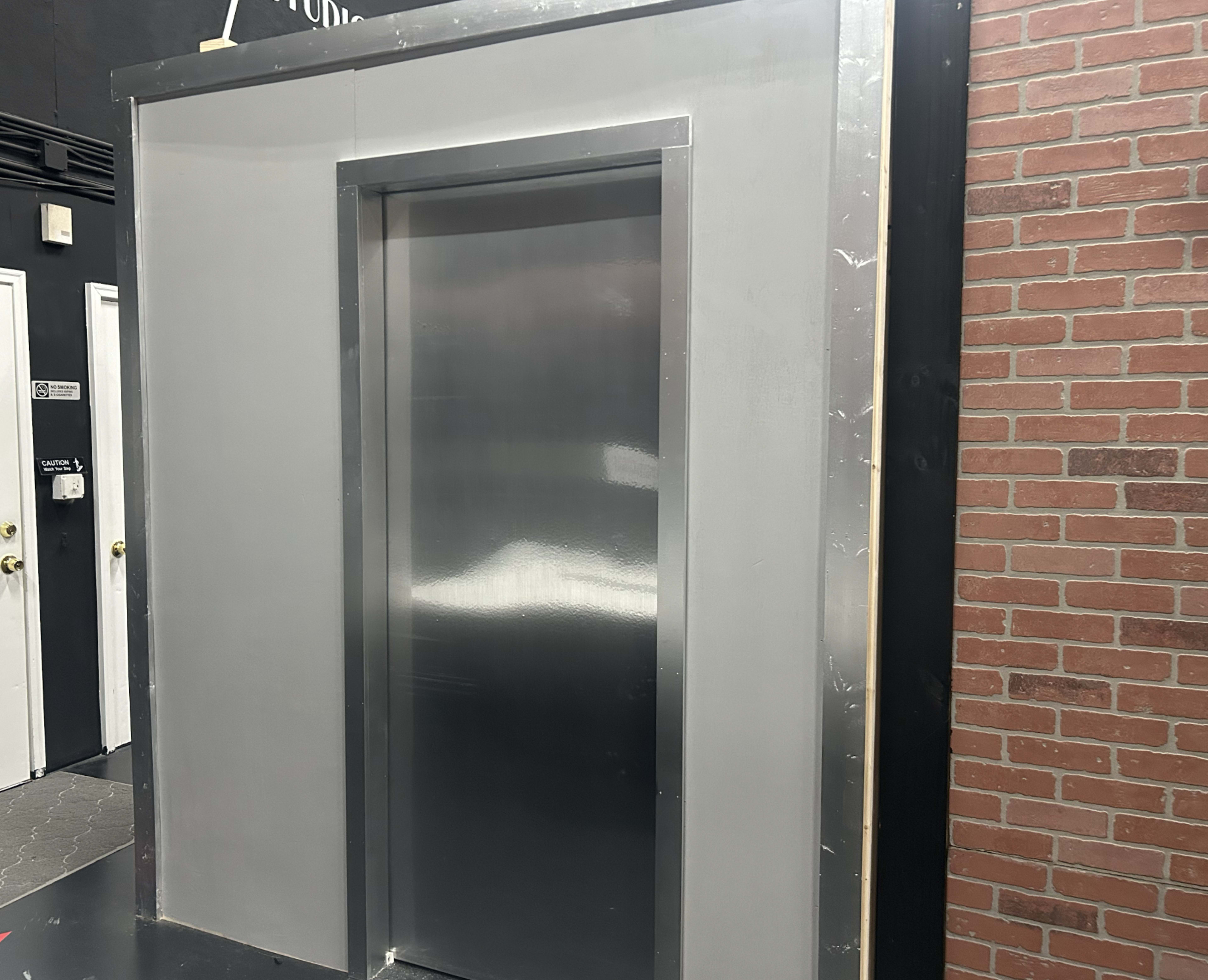 The image shows a metallic elevator door set within a gray enclosure against a brick wall backdrop.