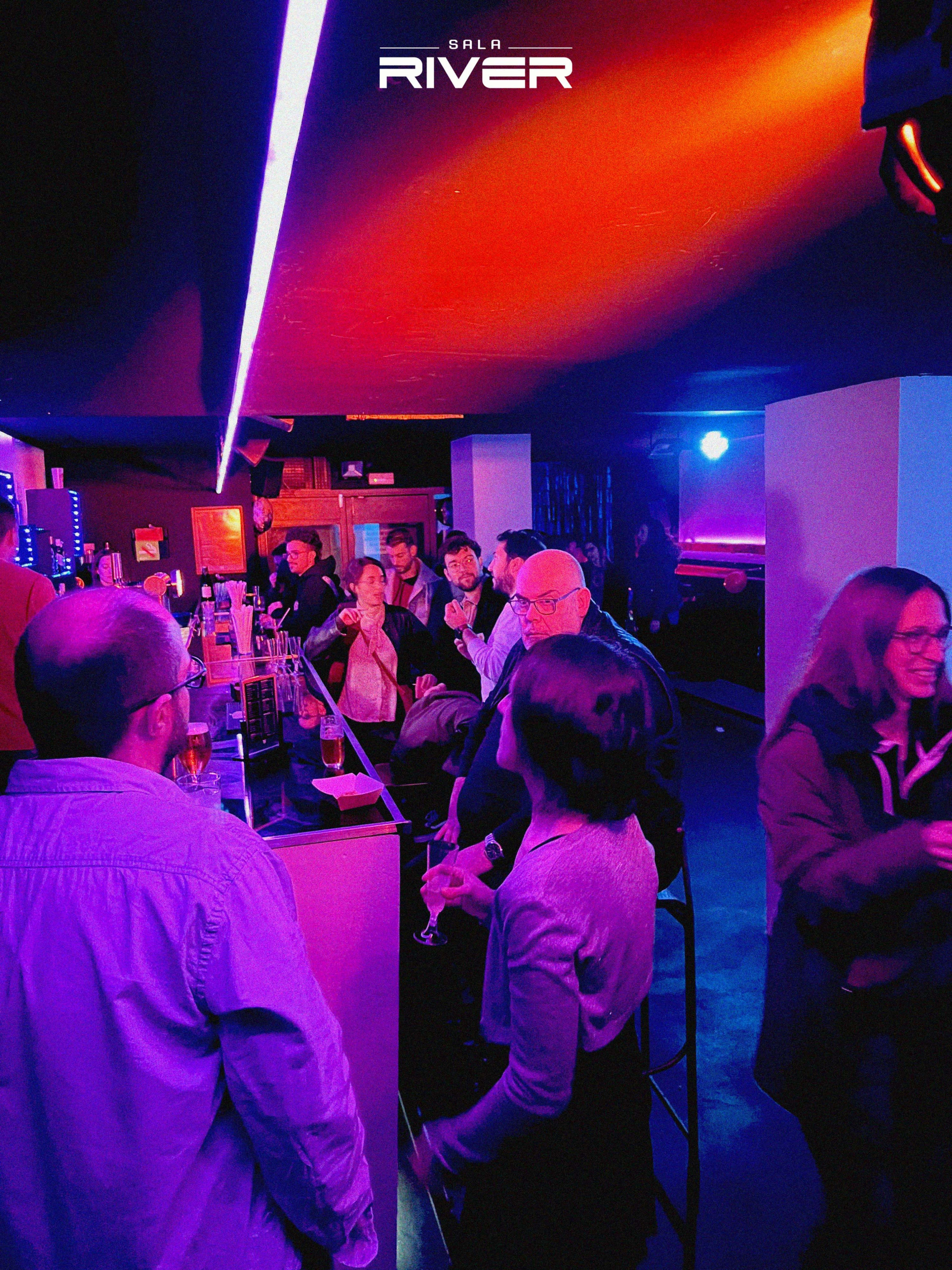 A lively bar scene with patrons socializing and drinks on the counter, illuminated by colorful lights.