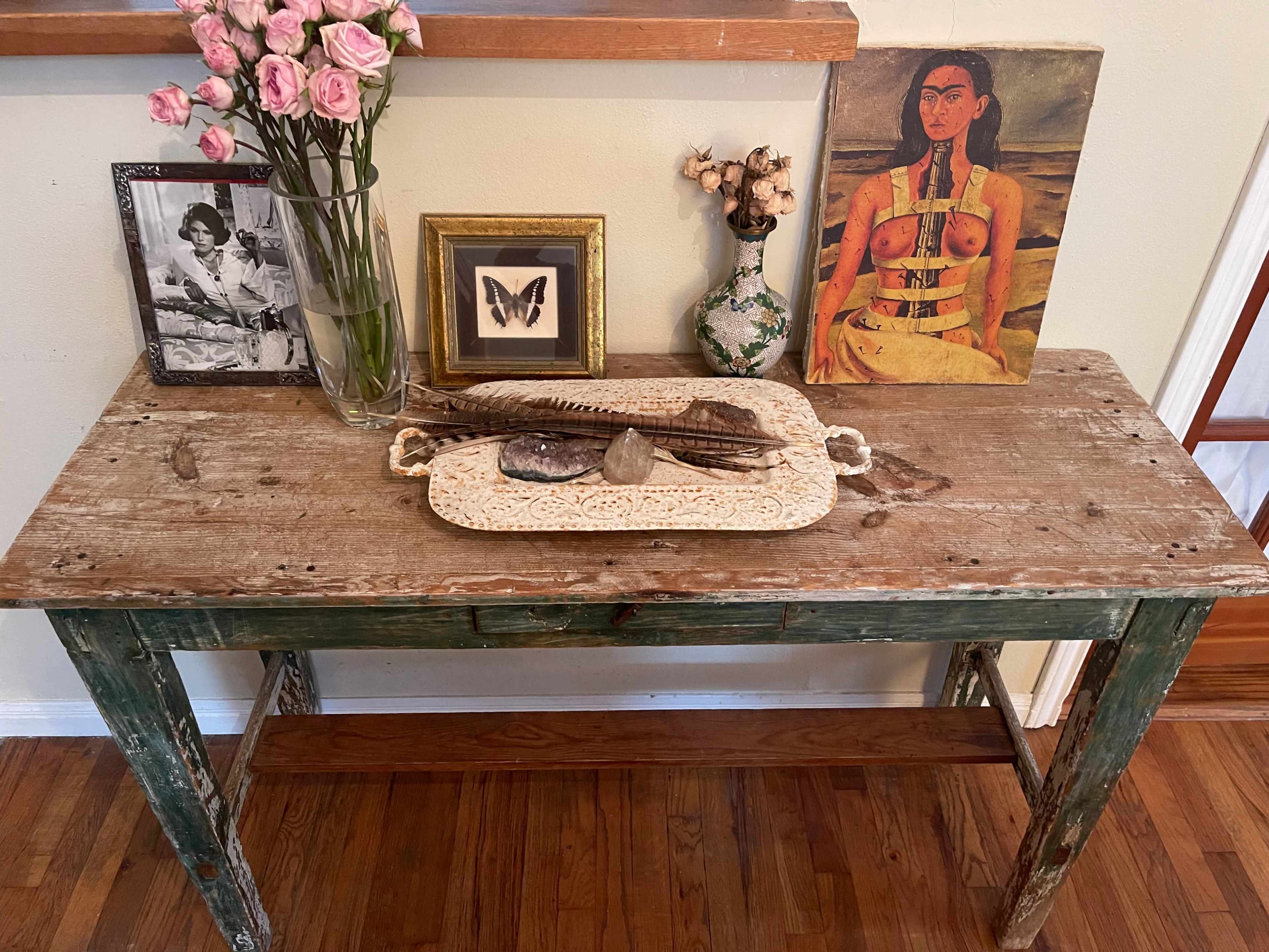 A rustic wooden table displays a vase of pink roses, framed photographs, a small art piece, and various decorative items arranged on a lace tray.