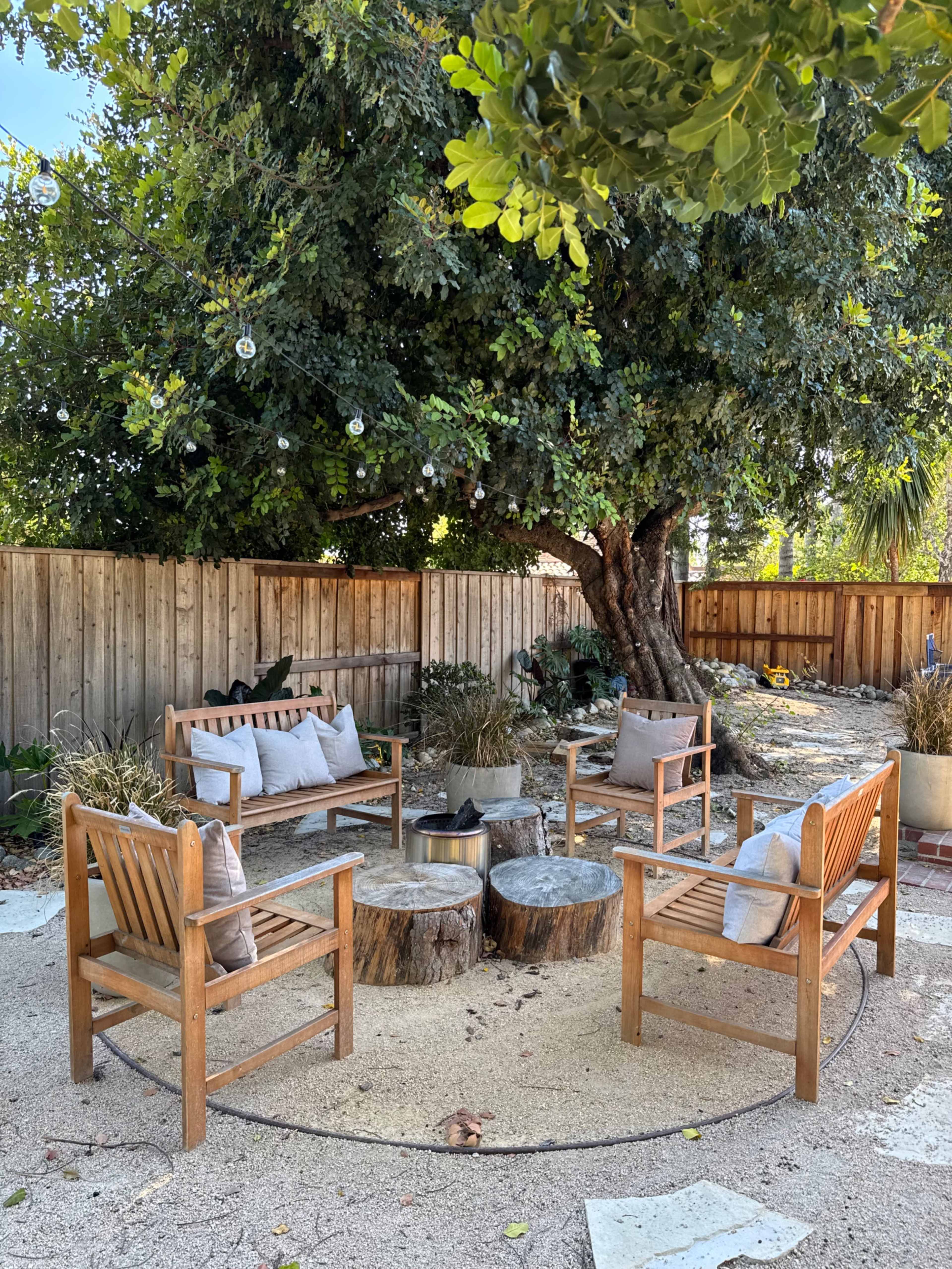 A circular seating area with wooden chairs surrounds a fire pit, located under a large tree in a fenced backyard.