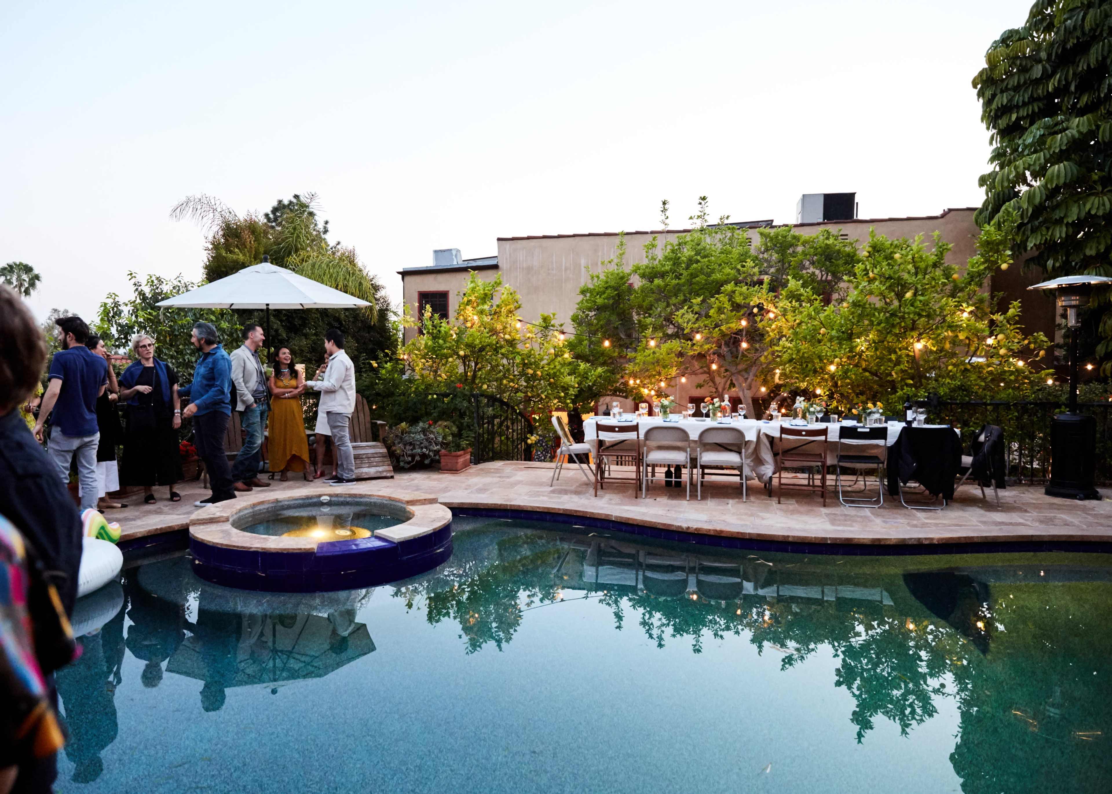 A gathering is taking place by a poolside area with tables set for dining and people socializing under string lights and shaded umbrellas.