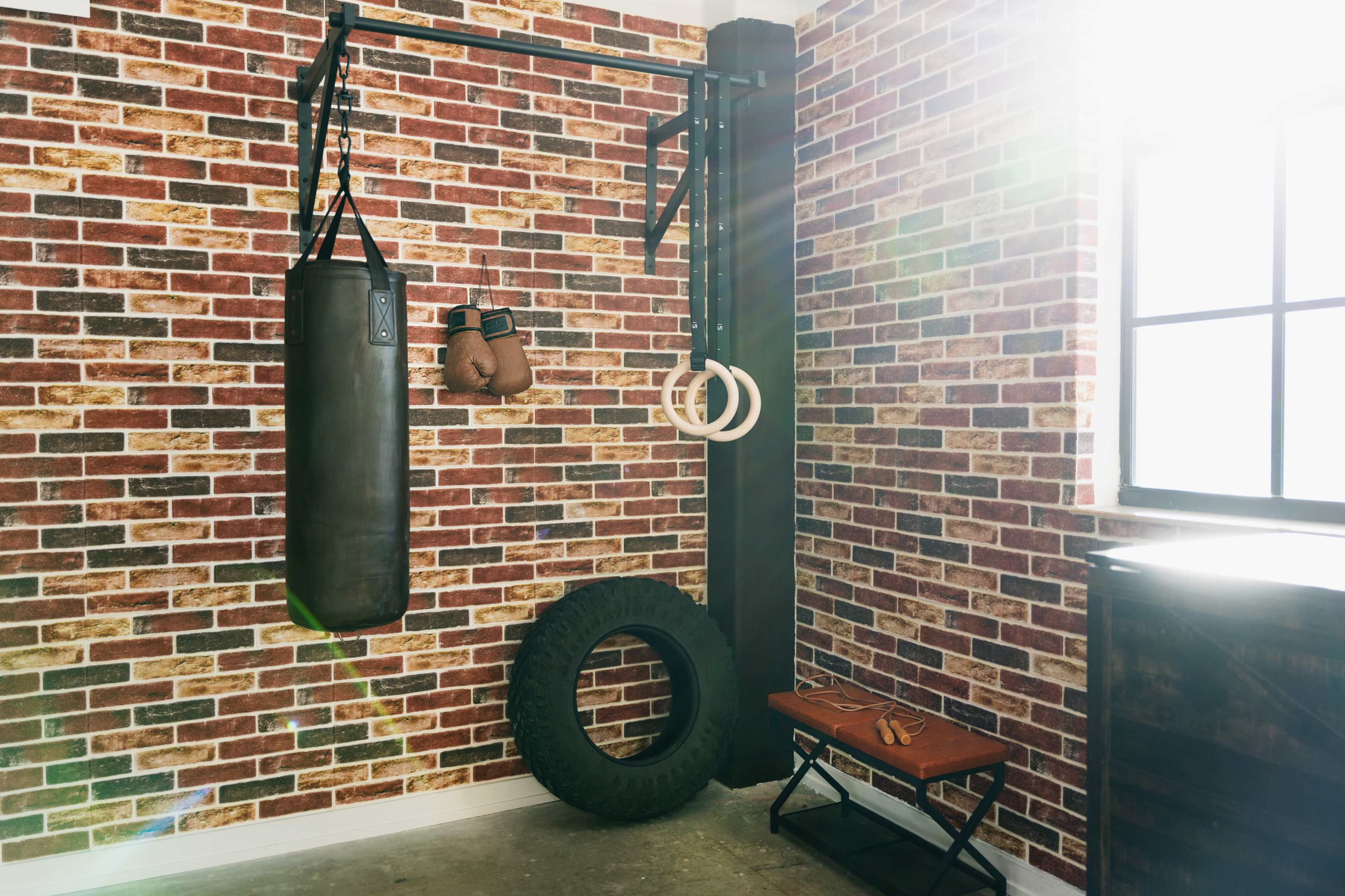 The image shows a home gym setup featuring a heavy punching bag, gymnastic rings, a tire, and a wooden bench against a brick wall.