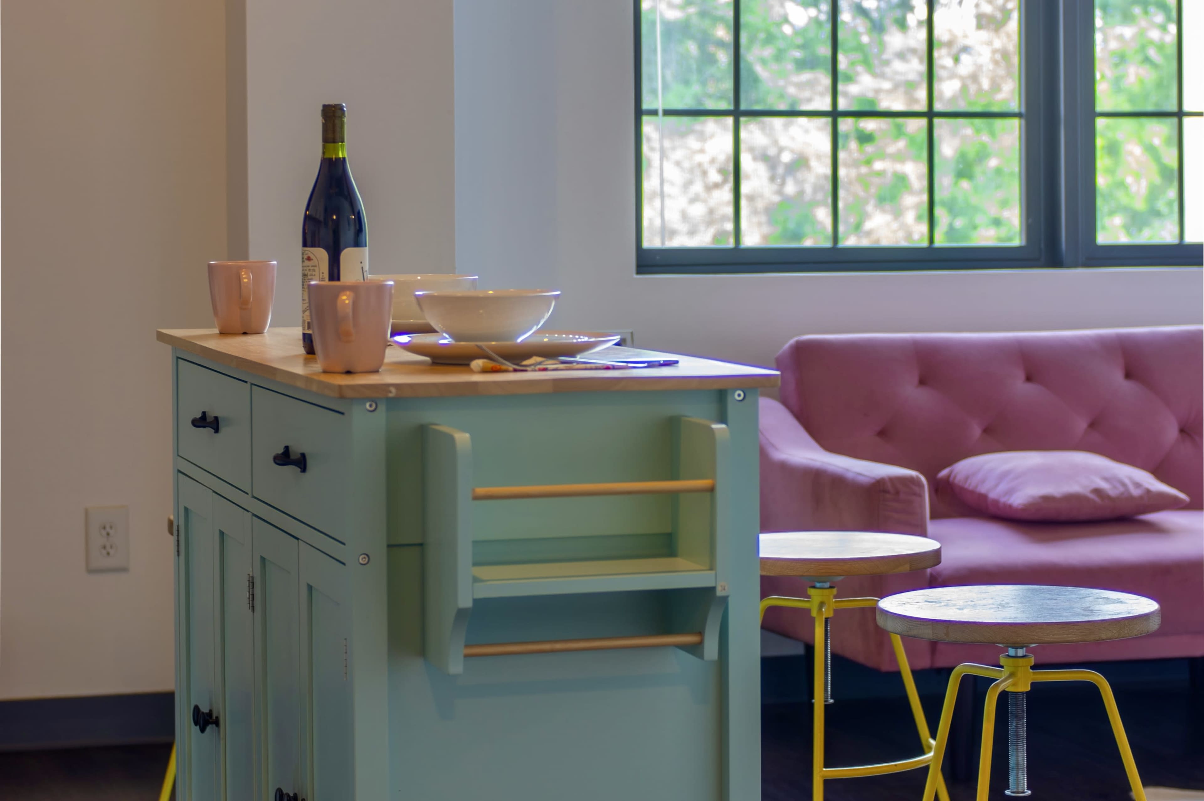 A pastel blue kitchen island with a wooden countertop and wine bottle sits near a pink tufted sofa and small round tables in a well-lit room with large windows.