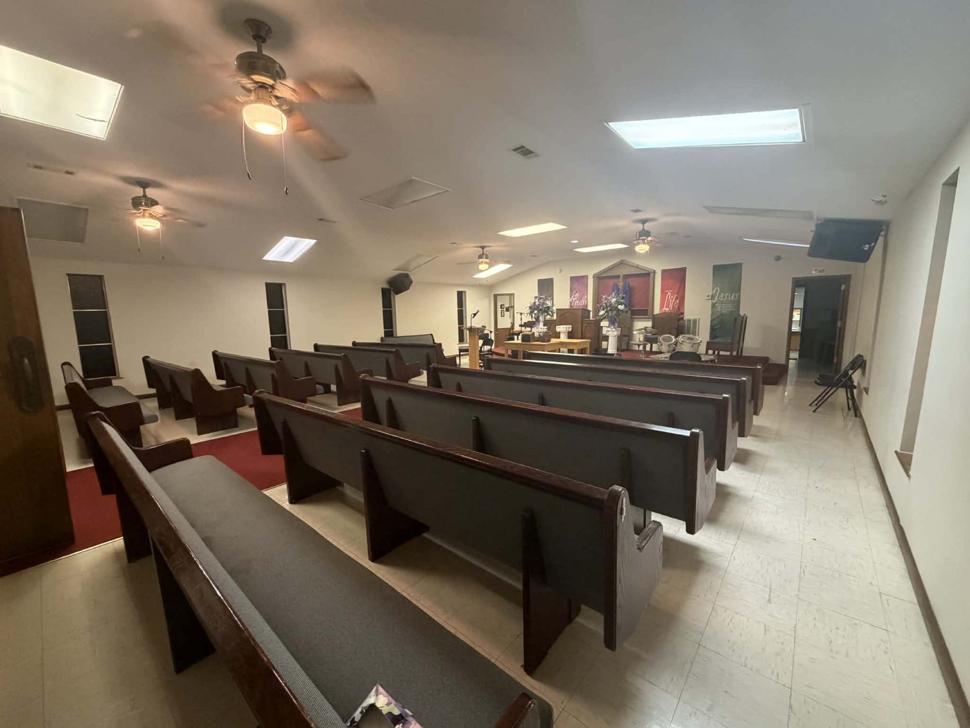 The image shows a quiet, empty church interior with wooden pews arranged facing a central altar area, illuminated by overhead lights.