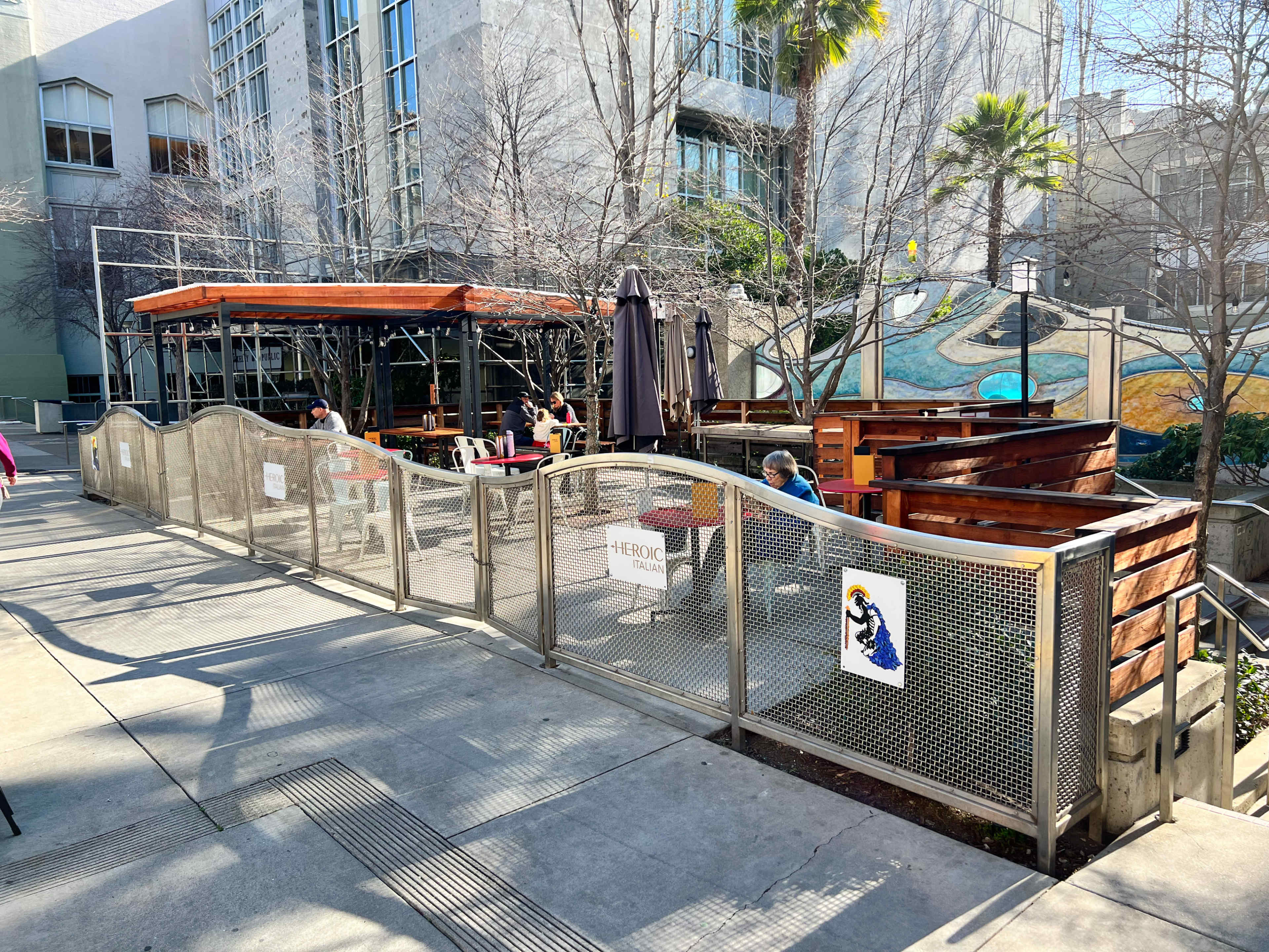 An outdoor dining area with wooden seating, shaded by umbrellas, enclosed by a metal fence, and set against a backdrop of urban buildings and greenery.
