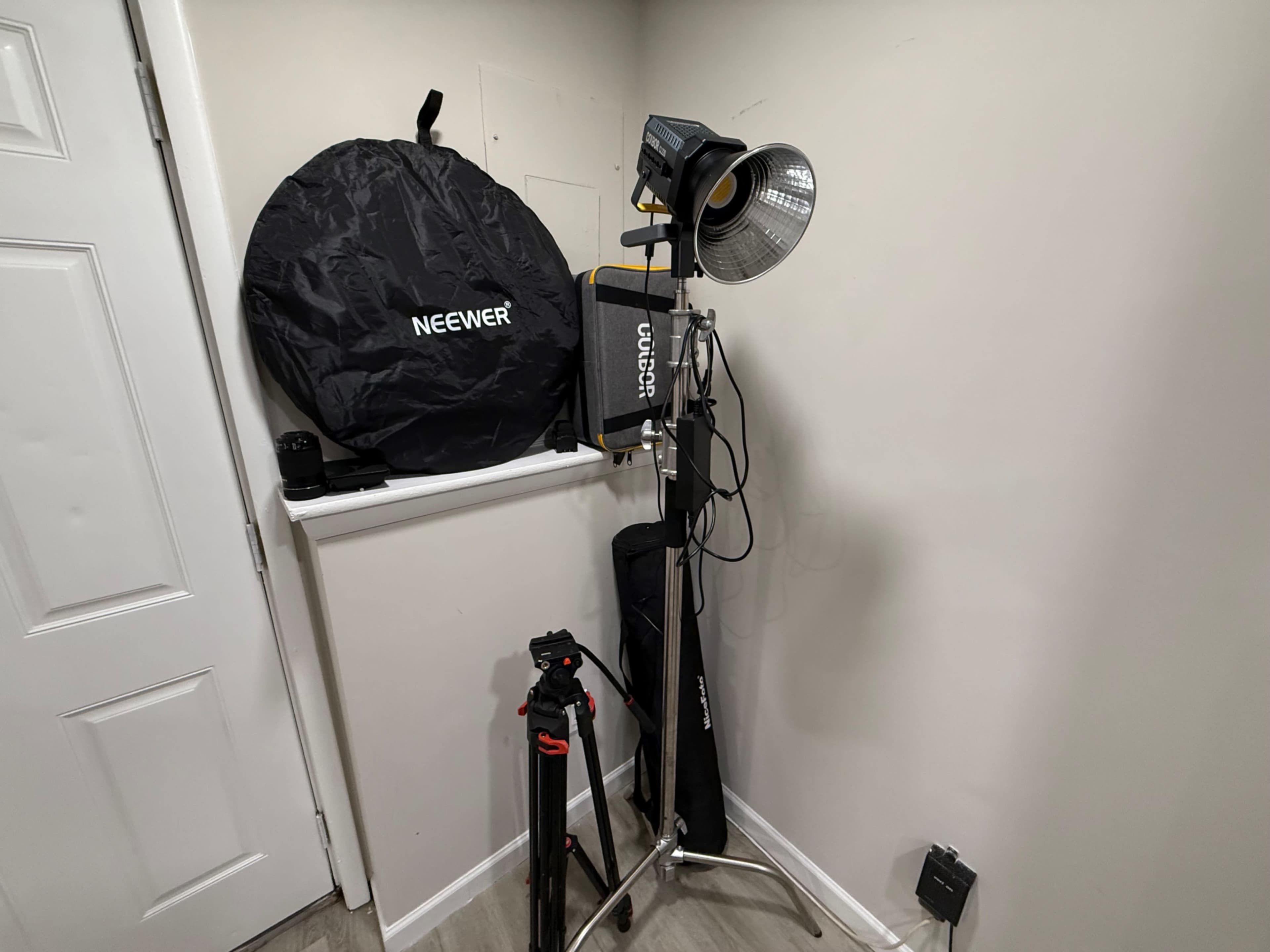 The image depicts a photography setup in a corner, featuring light equipment, a softbox, a tripod, and a power outlet.