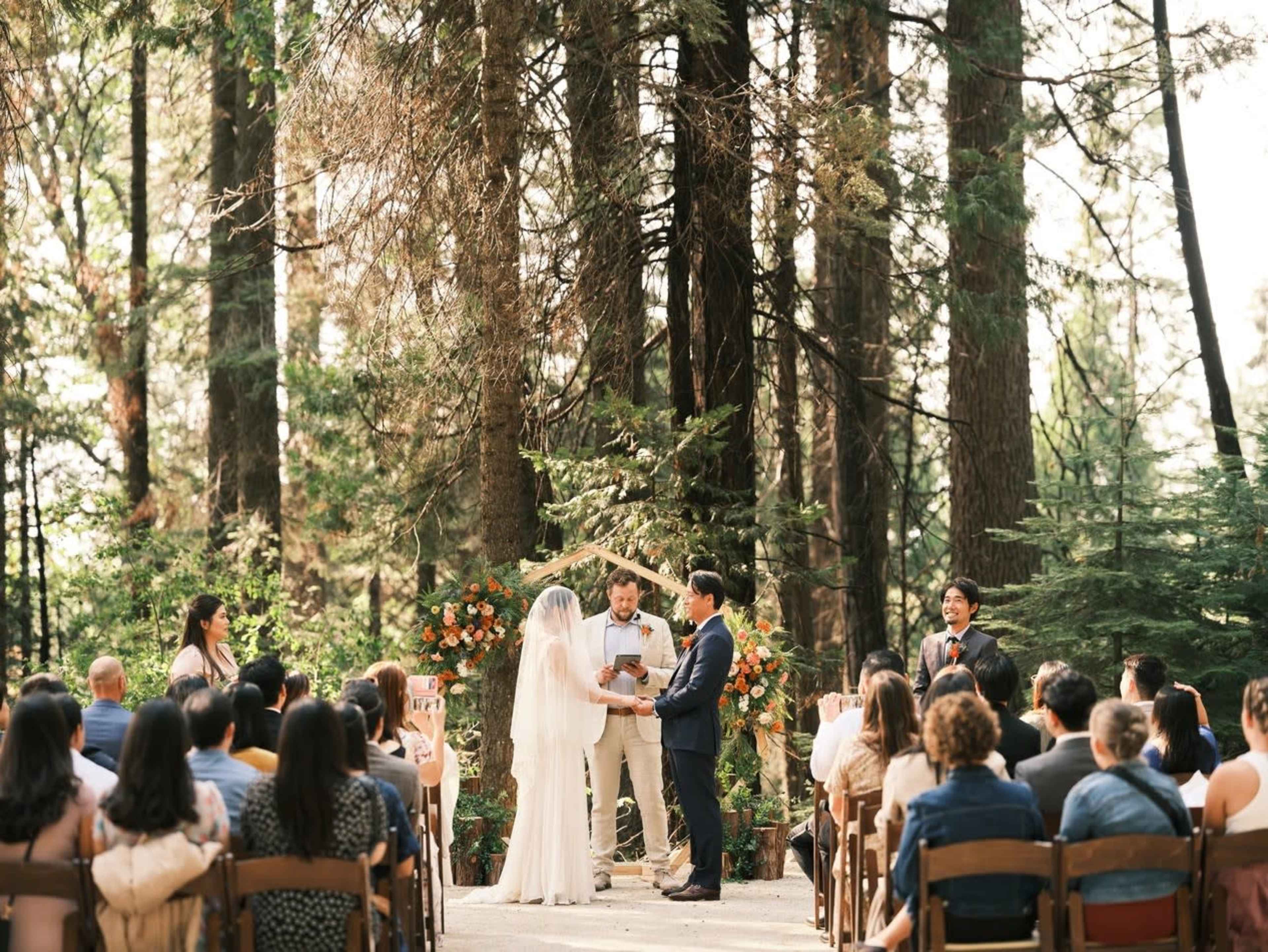 A couple stands under an arch adorned with flowers while exchanging vows in a forest setting, surrounded by guests seated on wooden chairs.