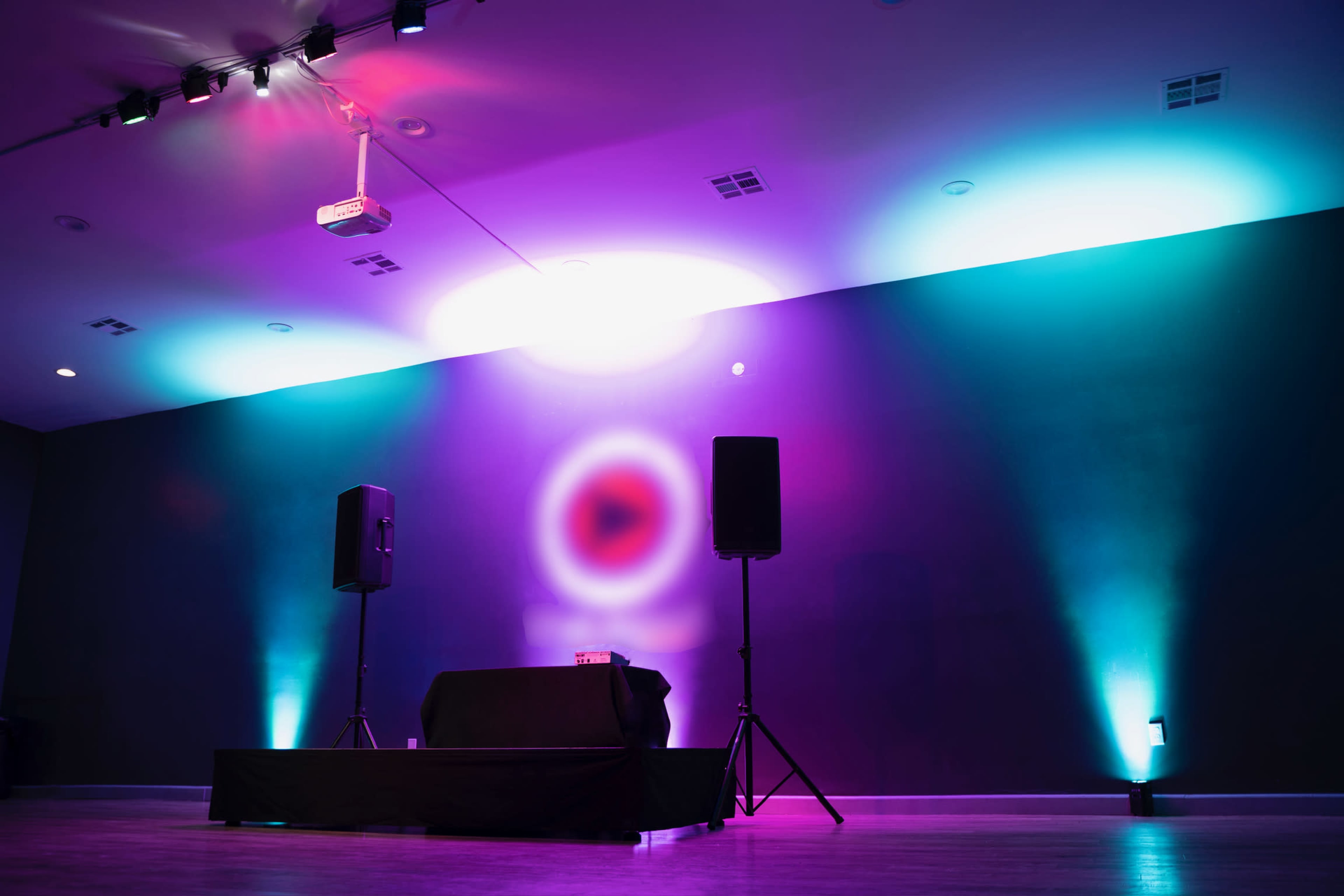 A stage setup with two speakers and a DJ table under colorful lighting in a dimly lit room.