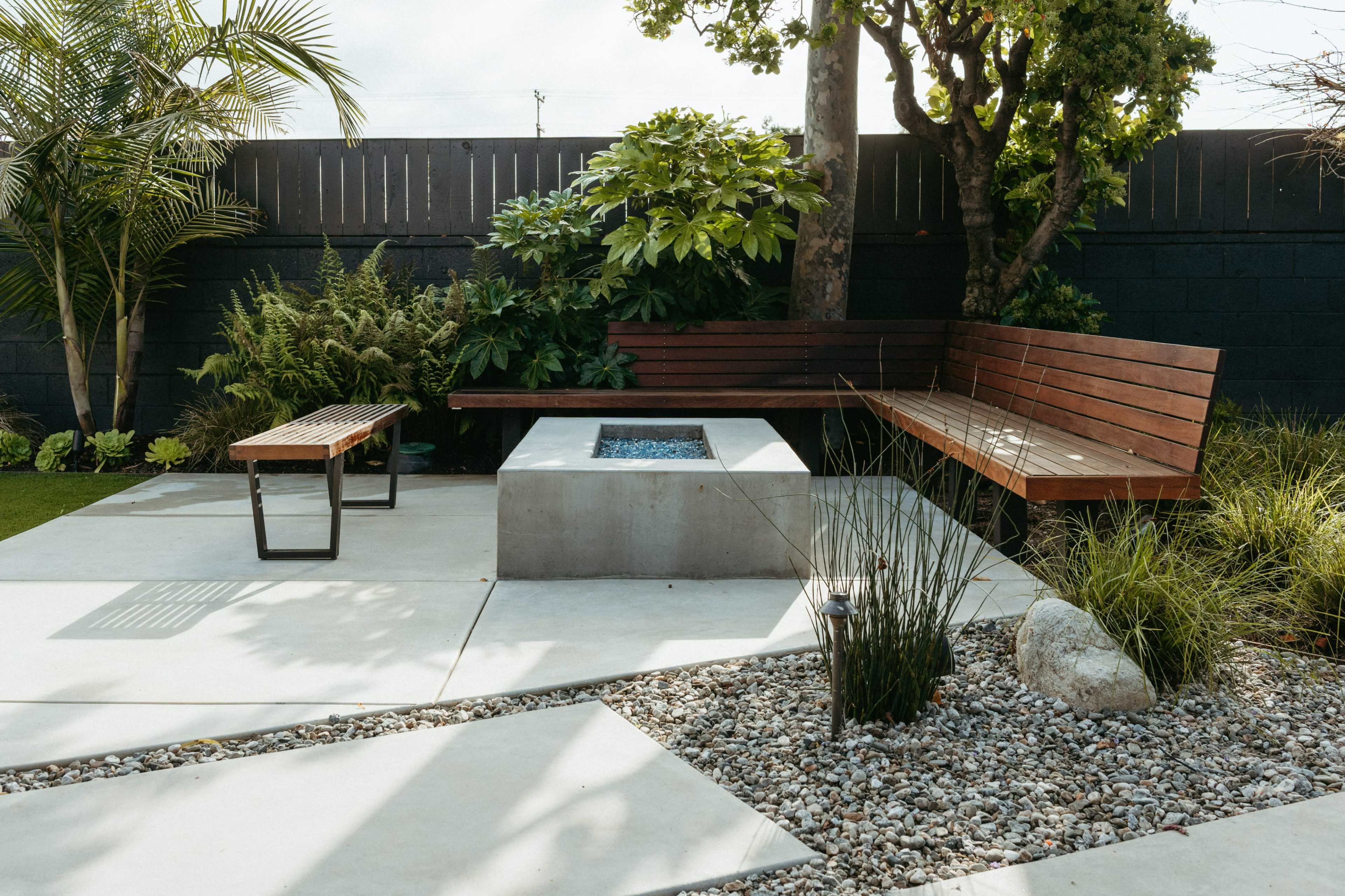 A modern outdoor seating area featuring a concrete fire pit surrounded by wooden benches, stone landscaping, and greenery.