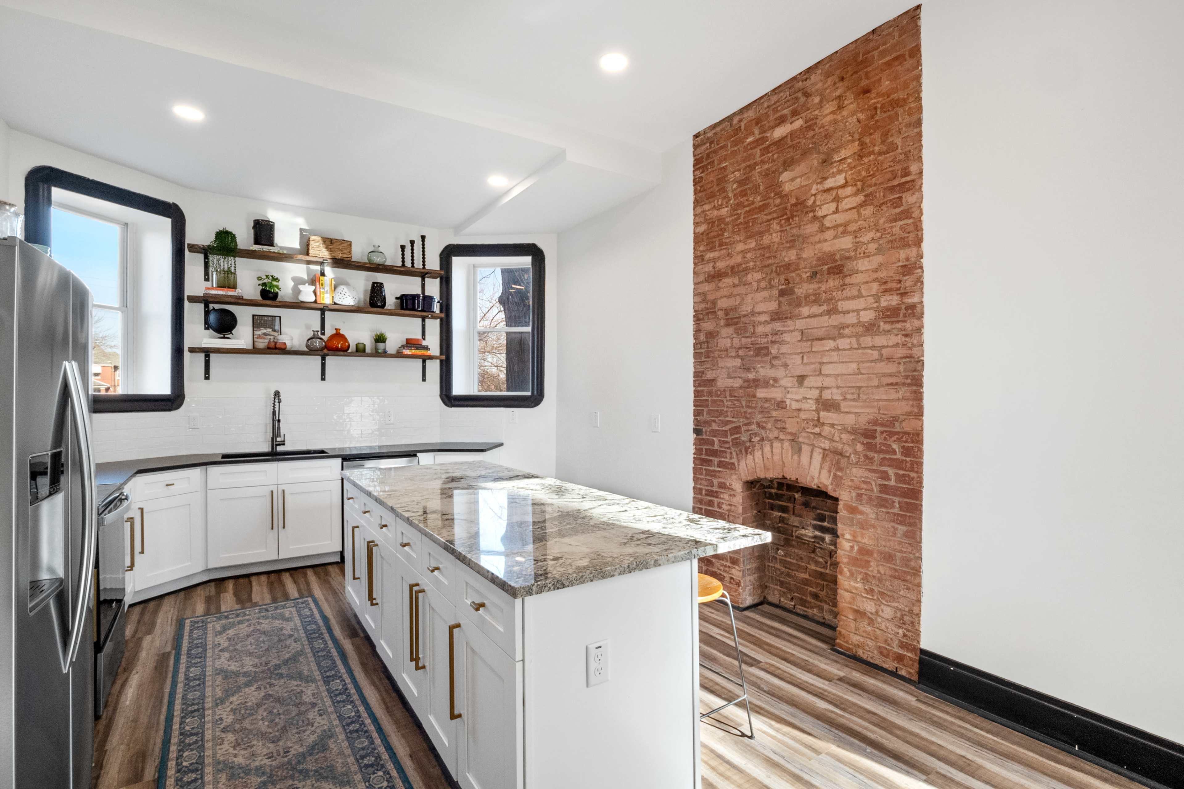 A modern kitchen features white cabinetry, a large island with granite countertops, and an exposed brick wall.