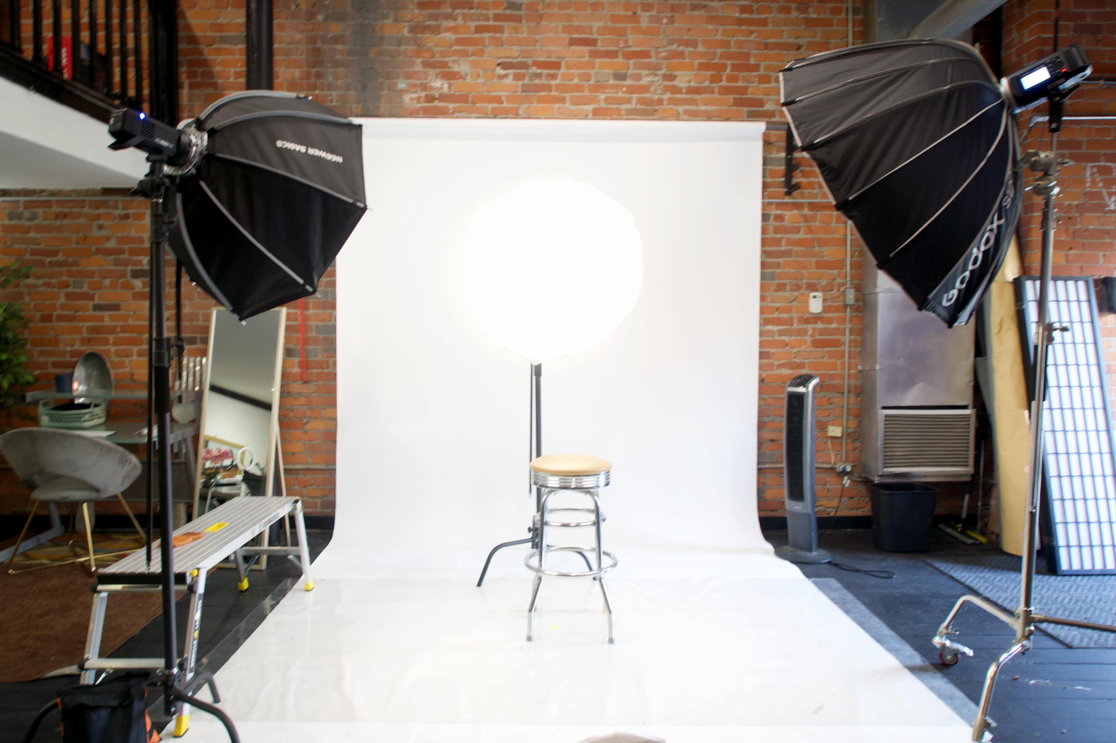 The image shows a photography studio setup with two large softbox lights illuminating a white backdrop and a single metal stool in the center.