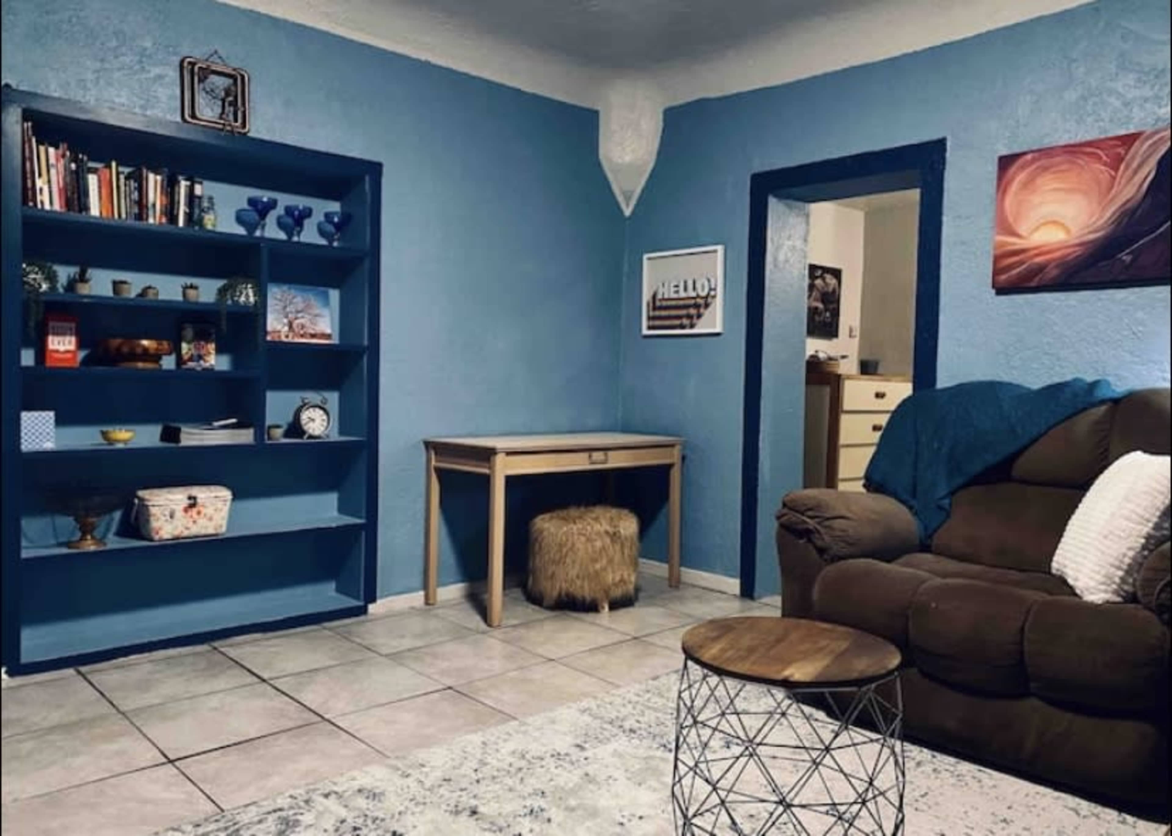 A living room with blue walls, a bookshelf filled with books and decorative items, a desk, and a brown couch.