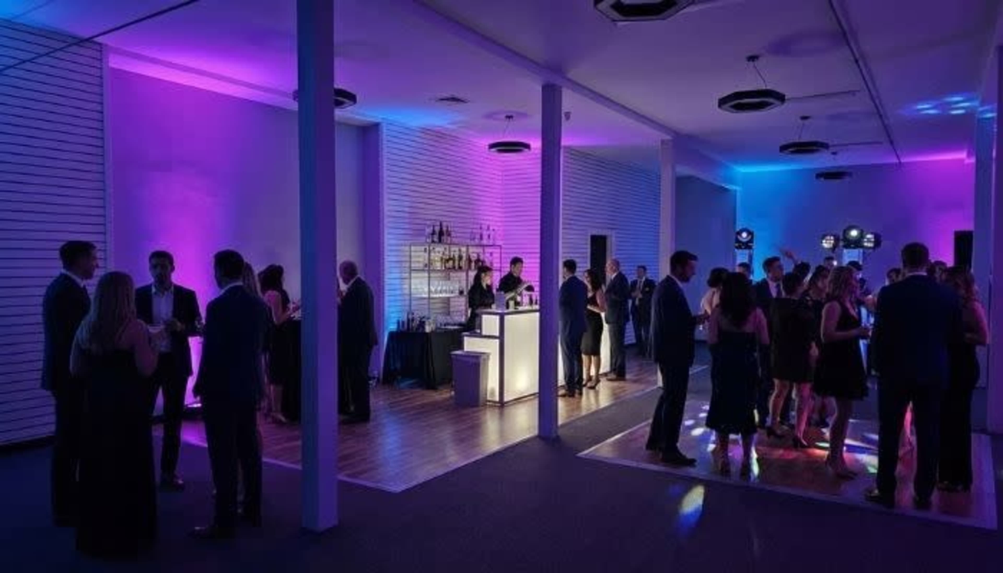 A lively event is taking place in a dimly lit room with purple and blue lighting, featuring a bar area and guests mingling on the dance floor.