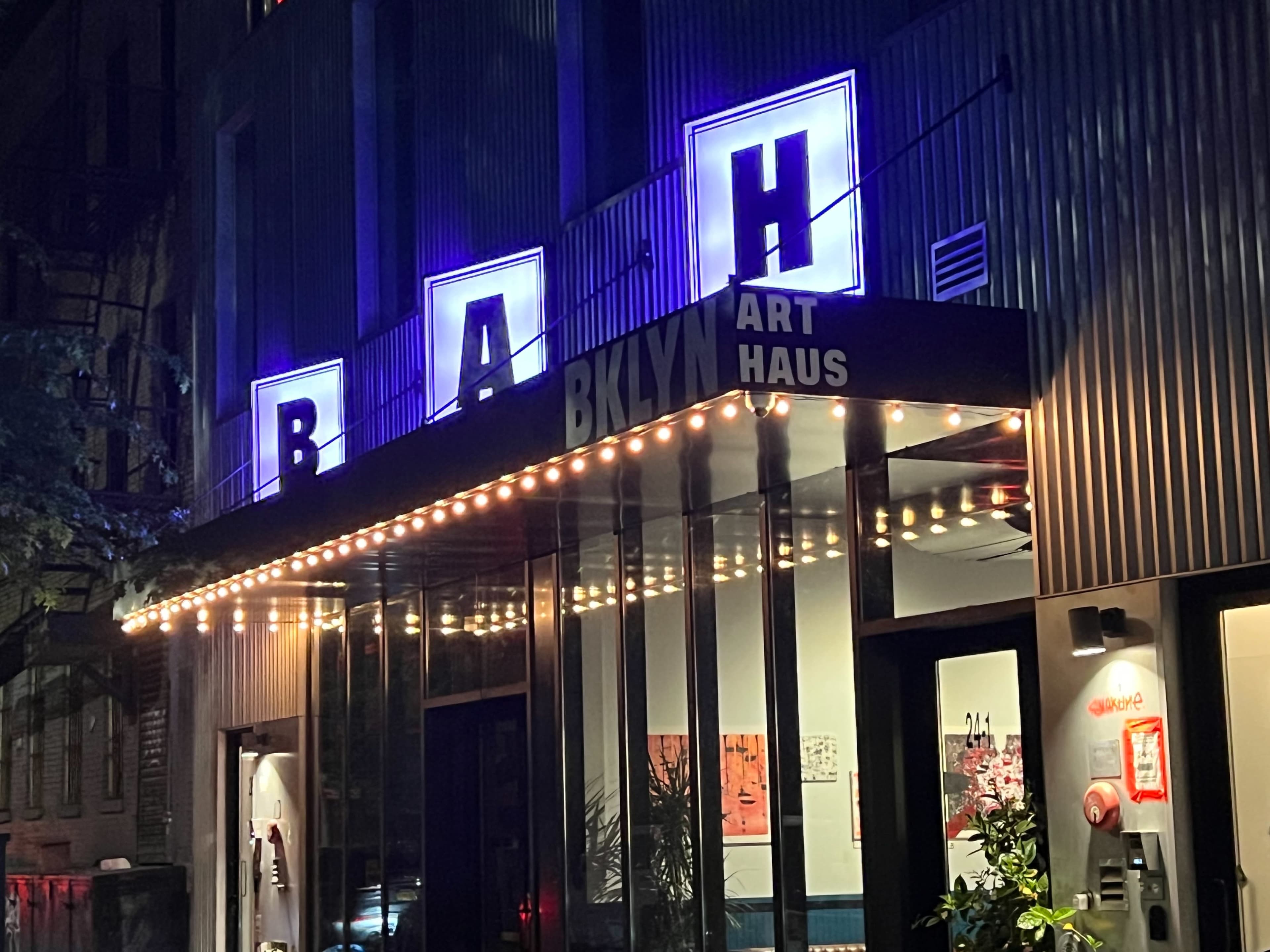 The image shows the illuminated entrance of BKLYN Art Haus, featuring large letters and string lights along the facade.