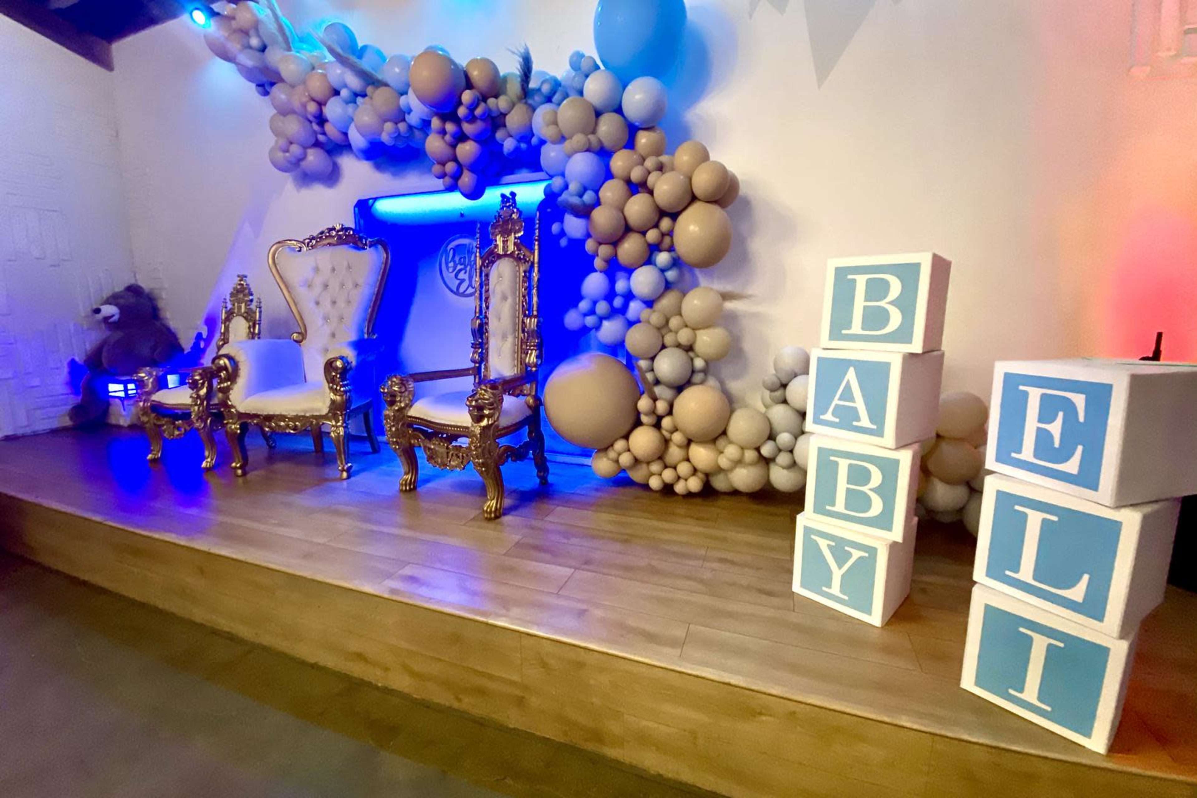 The image shows a decorated stage featuring two ornate chairs, a balloon arch in blue and beige tones, and large, colorful blocks spelling out "BABY ELI."
