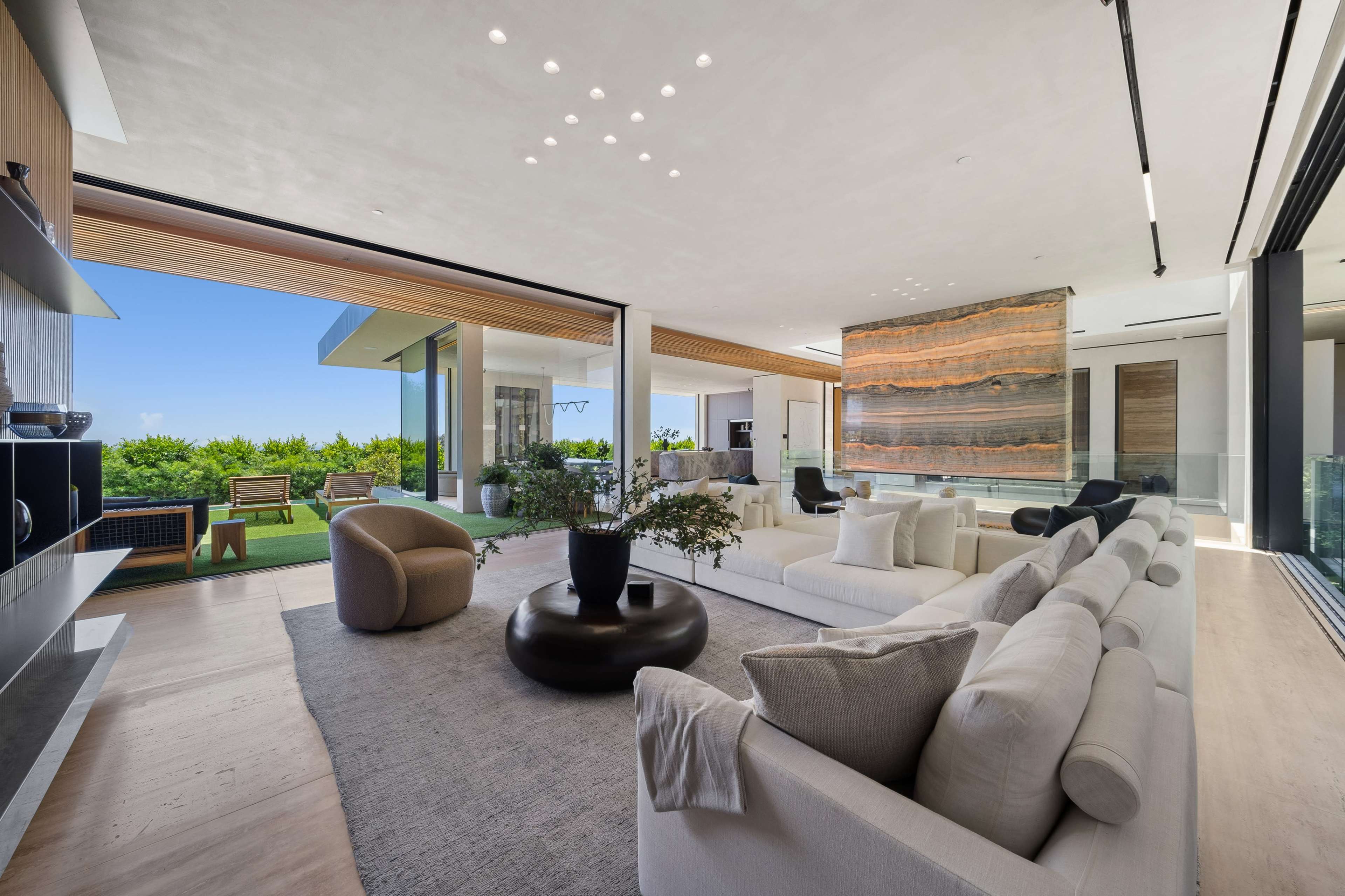 A modern living room with a light color palette, featuring a large sectional sofa, a round coffee table, and floor-to-ceiling windows that open to a view of greenery outside.
