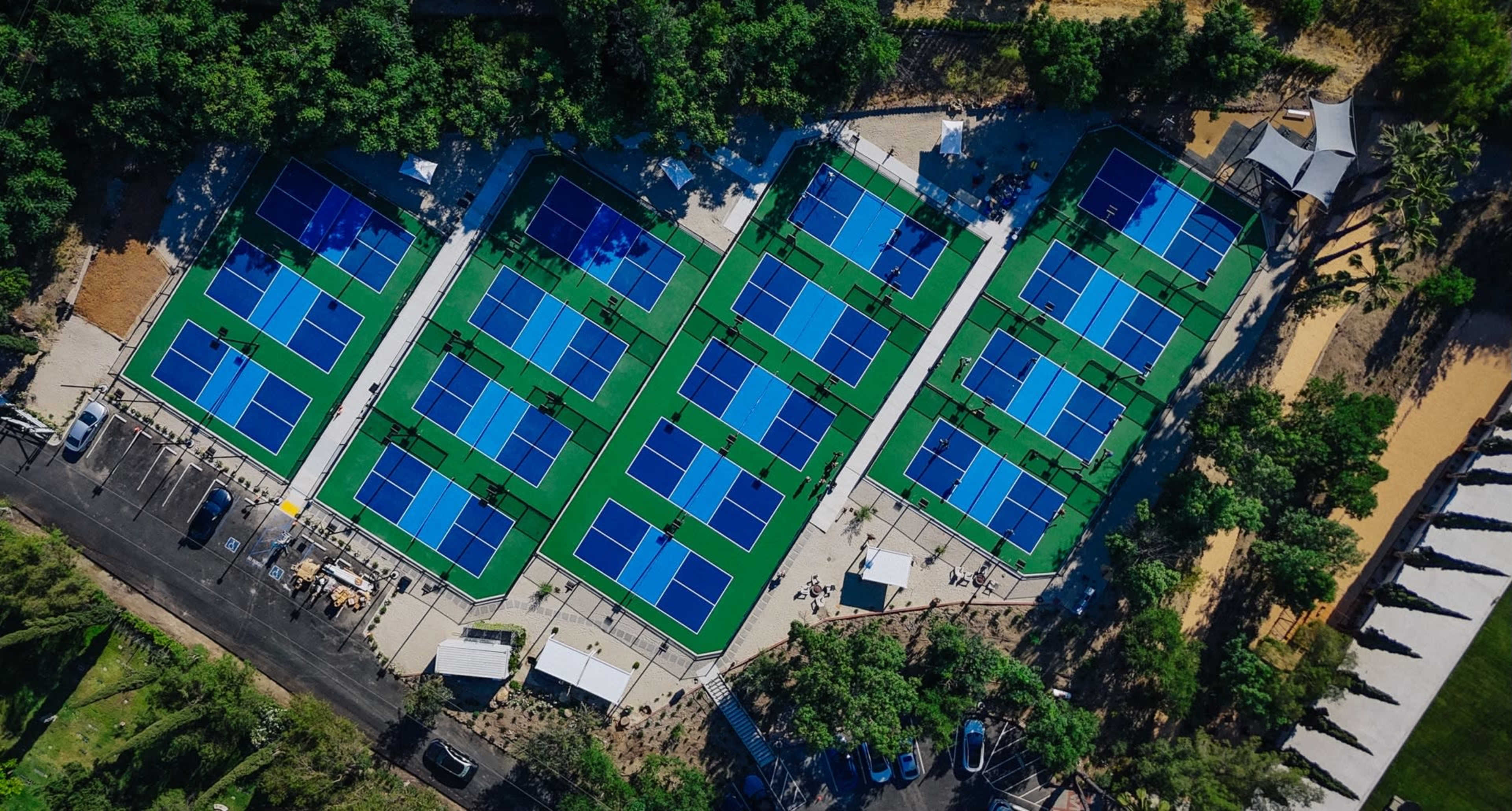 Aerial view of multiple tennis courts surrounded by trees and parked cars.