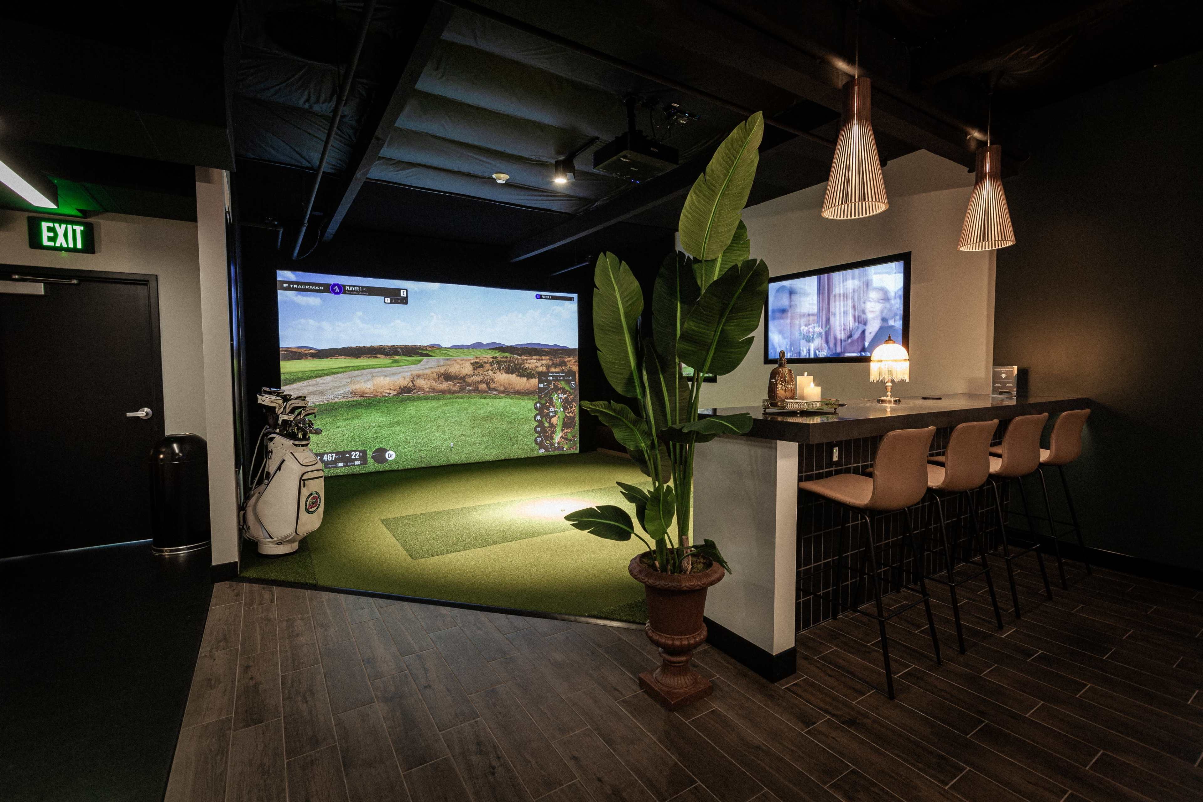 The image shows a modern indoor golf simulator space with a bar area, seating, and a large screen displaying a golf course.